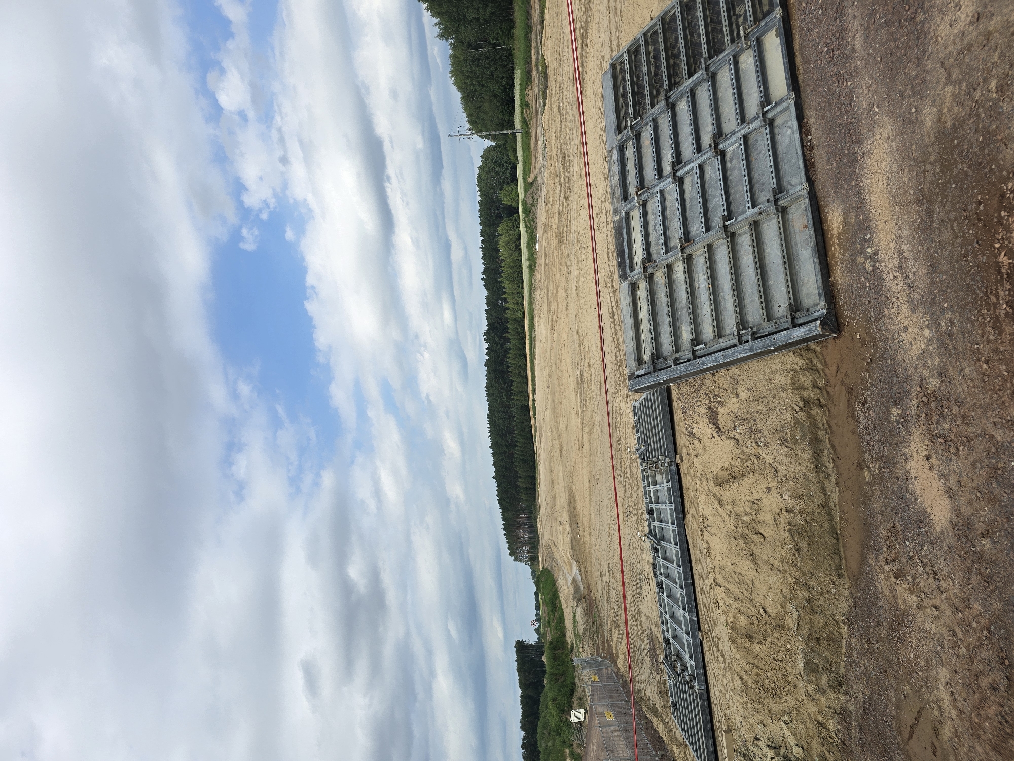 This is a landscape photograph taken on a cloudy day in Omulno, Poland. The foreground features a construction site with piles of sand and construction materials. Two large metal grates are laid out on the sand, likely for temporary road surfaces or ground support. A red caution tape is strung across the scene, indicating a restricted area. Beyond the construction site, there is a large expanse of cleared land, possibly for future development. In the distance, a dense line of trees forms a natural boundary, with a few scattered utility poles visible. The sky is filled with dramatic clouds, a mix of white and gray, with patches of blue sky peeking through. The overall impression is one of an ongoing construction project in a rural or semi-rural setting.