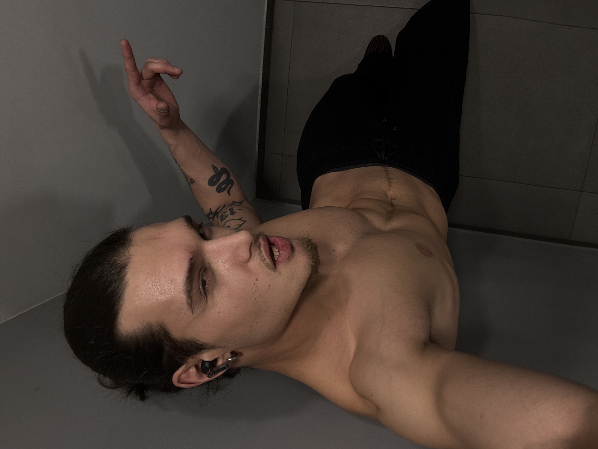 A man with long dark hair and a mustache lies on his back in the corner of a room with gray walls. He is wearing black pants and is shirtless, revealing his torso. He is pointing upwards with his right hand, which has tattoos on the forearm. He is wearing an earpiece in his left ear. The location is not discernible as being in Sofia, Bulgaria.