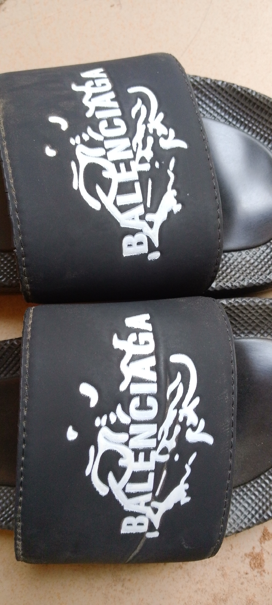 The image displays a close-up, overhead view of two black Balenciaga slides. The slides have a distinctive white graphic design and the Balenciaga logo printed on the straps. The straps are thick and appear to be made of a matte material, possibly rubber or synthetic leather, with white stitching along the edges. The white graphic on the straps is abstract and somewhat distressed, with the word "BALENCIAGA" in large, bold, white capital letters incorporated into the design. Some of the letters are partially obscured by the graphic elements, giving it a stylized, graffiti-like appearance. The soles of the slides are also black, with a textured pattern on the outer edge, and a smooth inner surface where the foot would rest. The slides are lying flat on a light-colored, textured surface that appears to be concrete or a similar ground material, possibly outdoors. The lighting suggests it might be daytime, with natural light illuminating the subjects. The setting appears to be casual, and there are no people visible in the image. The overall impression is a product shot, focusing on the design and branding of the slides. The context provided indicates the location is Jalingo, Nigeria.