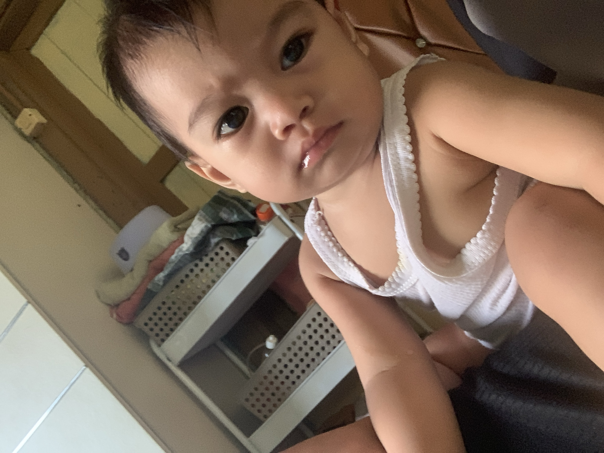 The media file is a close-up, angled selfie of a baby, likely taken indoors. The baby has dark hair and is looking directly at the camera with a serious expression. They are wearing a white ribbed tank top. In the background, to the left of the baby, is a light-colored wall and a wooden frame. Below this, a multi-tiered plastic storage unit is visible, filled with folded clothes in muted colors, including pink, cream, and a green and white striped pattern. A white, possibly ceramic or plastic, item is also visible on the top shelf of the storage unit. To the right of the baby, a dark fabric, possibly a couch or blanket, is visible. The lighting suggests it could be daytime. No text is visible in the image. The location context provided is Sungai Meriam, Indonesia.