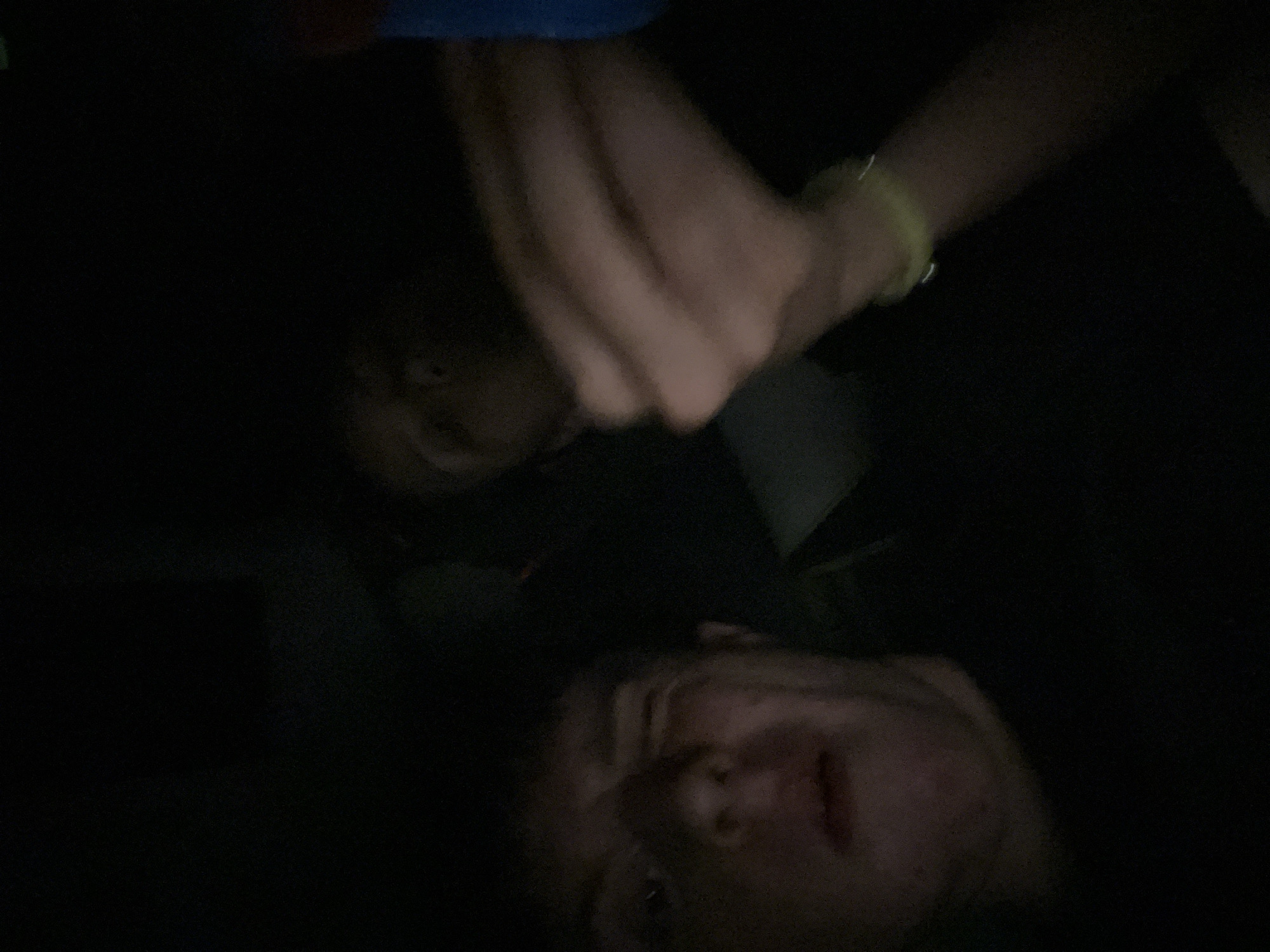 Two people are visible in the dark image. One person is lying down, facing upwards with their face visible at the bottom of the frame. The other person is above them with a hand raised, a wristband is visible on their wrist, also face visible. The setting appears to be indoors in dim or no light. The location of Popovitsa, Bulgaria is not evident from the image. No specific actions or interactions can be definitively determined.