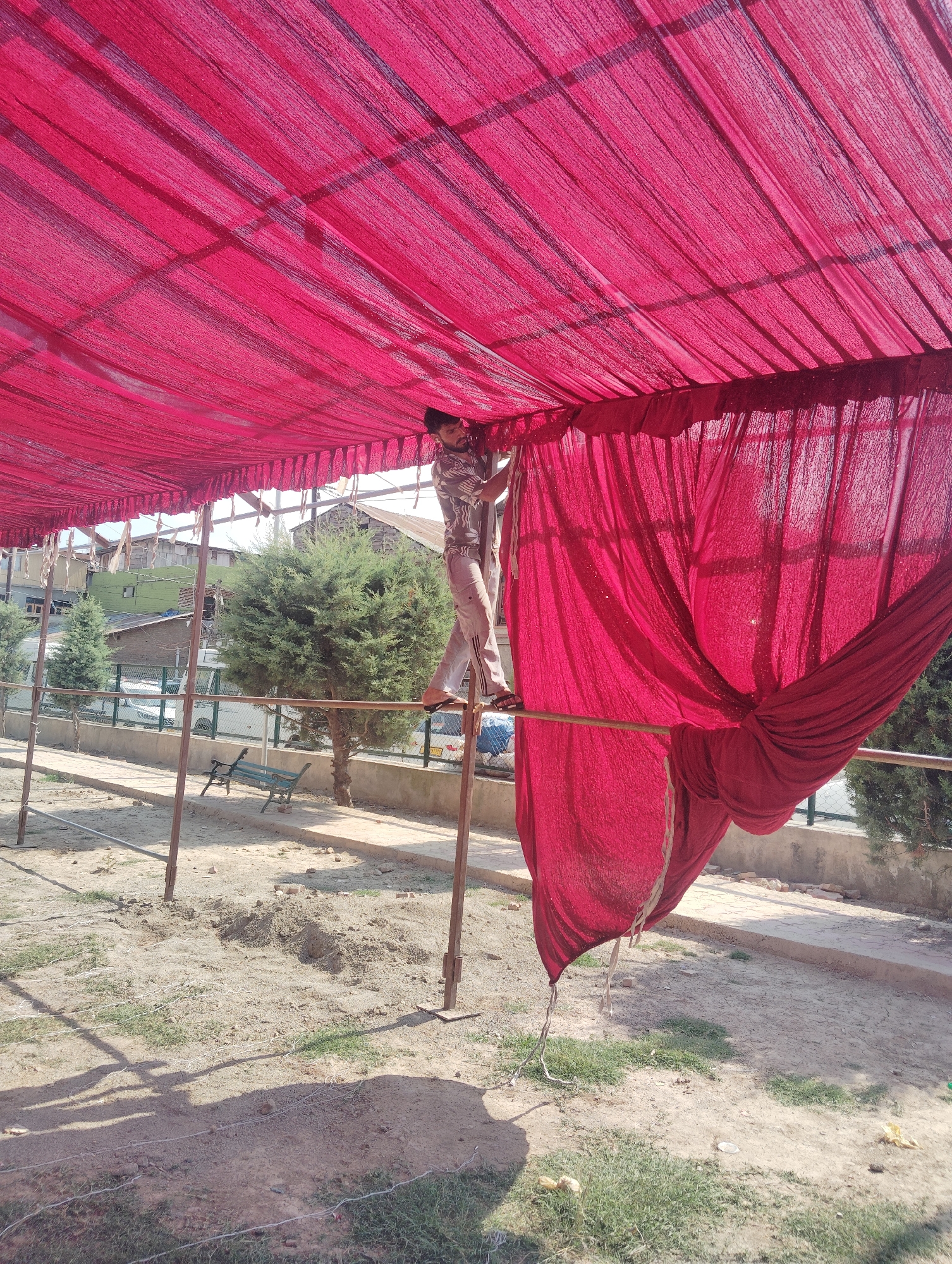 A person is standing on a metal pole, adjusting a large red fabric canopy outdoors. The canopy is supported by metal poles and appears to be made of a sheer, textured material. The person is wearing a patterned shirt and light-colored pants, with their feet in sandals. They are positioned on the right side of the image, reaching up towards the canopy. In the background, there are trees, buildings with various colored roofs, a fence, and a bench. The ground is mostly dry dirt with patches of grass. The scene appears to be in an open, outdoor area, possibly a park or public space in Srinagar, India, given the architectural style of the buildings and the vegetation. The lighting suggests it is daytime.