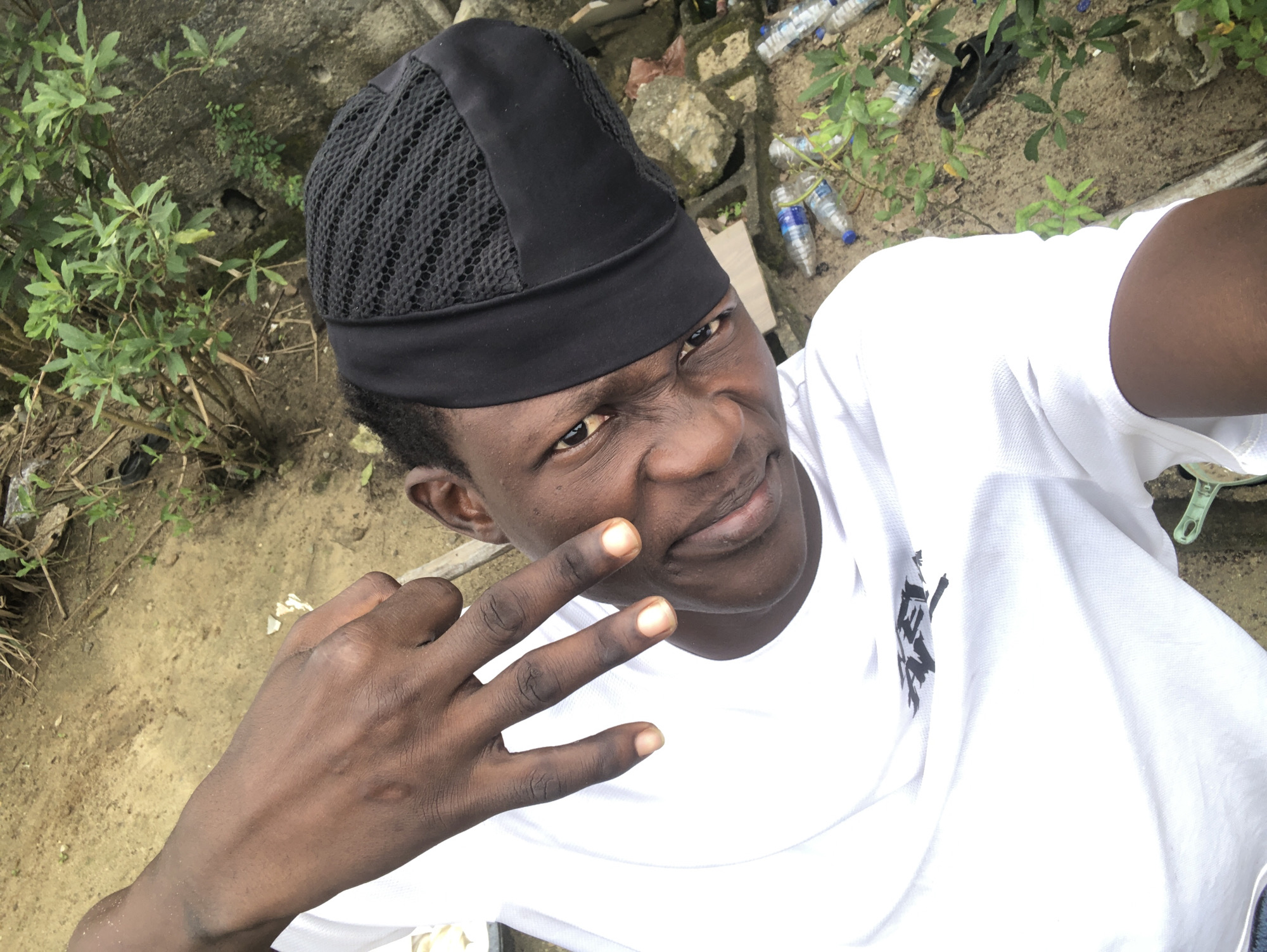 In Ado, Nigeria, an outdoor scene depicts a young Black male, facing upwards and to the left, with his head tilted. He wears a white t-shirt with a black logo and a black, textured cap. His right hand is raised, with two fingers extended in a V-sign. The background consists of dry, sandy ground, some foliage, rocks, and discarded plastic water bottles, suggesting an urban or semi-urban environment.