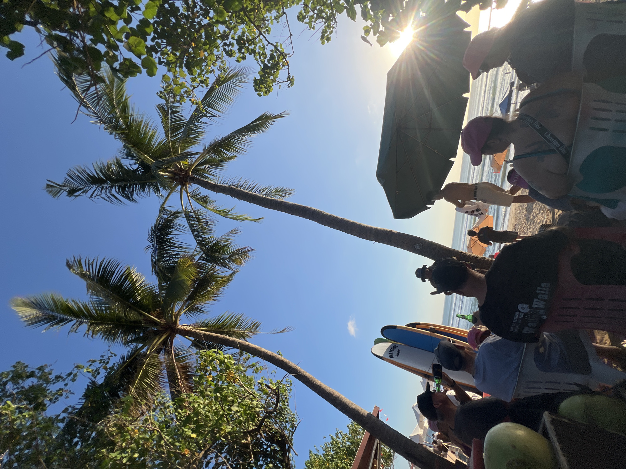 This image depicts a beach scene in Kuta, Indonesia, with several people seated and standing. Two prominent palm trees frame the upper portion of the image, with their fronds reaching towards a clear blue sky. In the foreground, several people are seated, some with drinks in hand. To their left, a collection of surfboards leans against each other. Behind the seated individuals, the beach stretches out towards the ocean, with waves visible in the distance. Sunlight streams in from the right, creating a bright lens flare and casting a warm glow on the scene. The overall atmosphere suggests a relaxed, beachfront setting, likely a bar or cafe, with patrons enjoying the late afternoon or early evening.