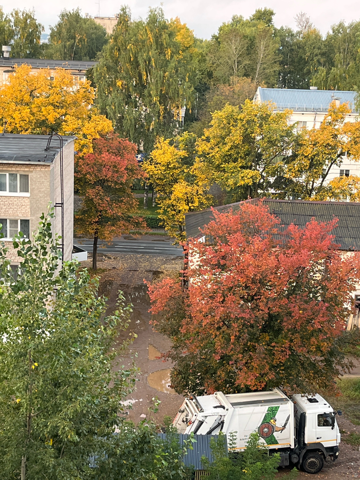 In Chaikovsky, Russia, this overhead perspective captures an autumn scene dominated by residential buildings and vibrant foliage. The foreground features a white garbage truck parked on a dirt and asphalt area, partially obscured by green trees. The truck displays a circular emblem with a warrior figure, a sword, and a shield. Beyond the truck, a building with a corrugated roof stands, with a large tree displaying fiery red and orange leaves directly in front of it. To the left, a multi-story brick building with visible windows and a dark roof is present, partially covered by lush green foliage. Further in the background, more buildings with light-colored facades and blue and white roofs are visible, interspersed with trees showcasing a spectrum of yellow and green leaves, indicating the fall season. The ground in the mid-ground appears wet with puddles.