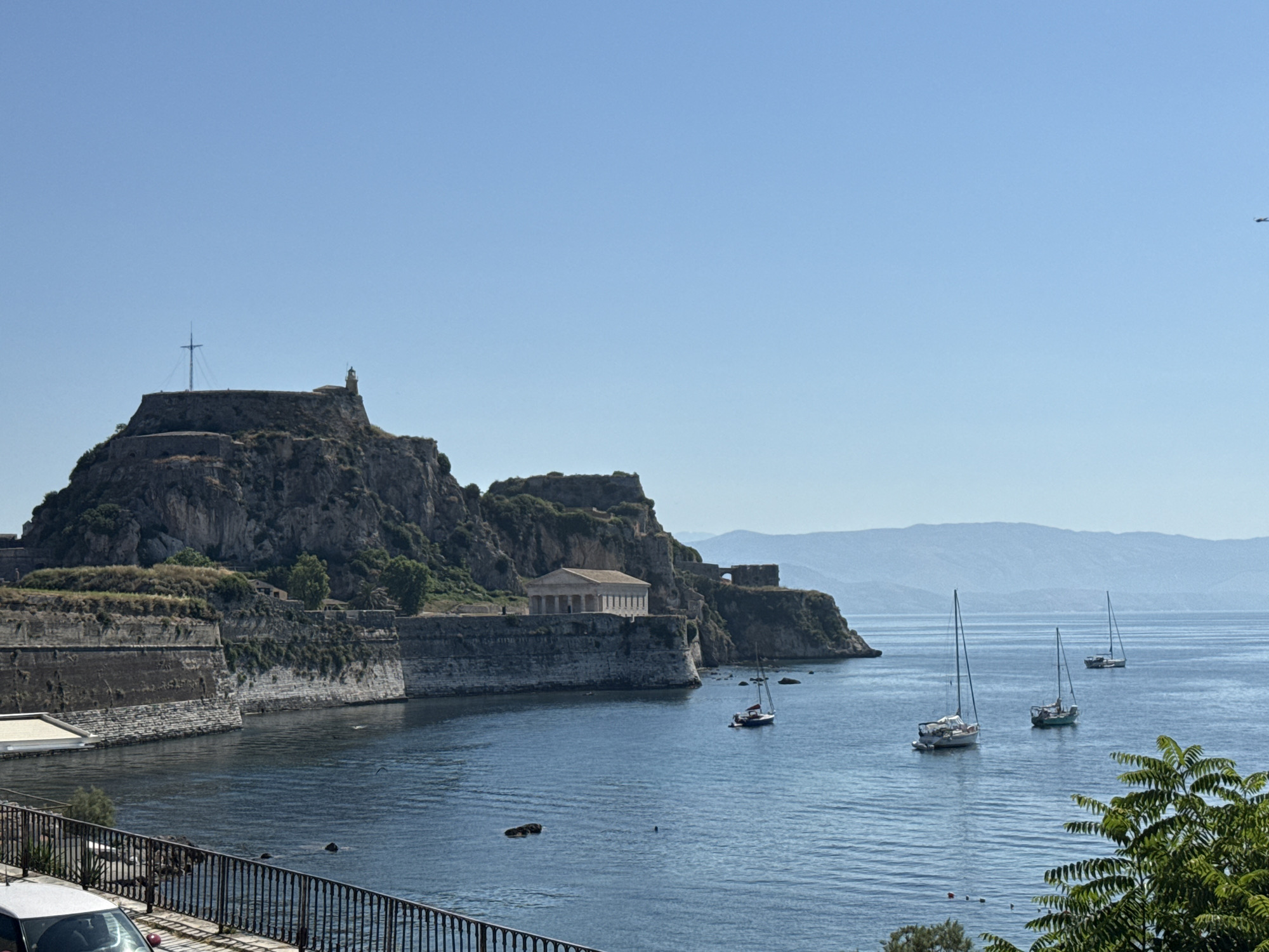The image depicts a scenic view of the Old Fortress of Corfu, Greece, from a slightly elevated perspective. In the foreground, a black metal railing separates the viewer from a paved area with a white car visible on the left. The sea occupies the middle ground, with several sailboats anchored or cruising. Some dark, indistinct shapes, possibly buoys or submerged objects, are visible in the water.
The main subject of the image is the Old Fortress, a historical fortification built on a rocky promontory. Its stone walls and ramparts are prominent, with vegetation growing on some sections. A building with classical columns, likely a church or administrative building, is situated within the fortress walls. A lighthouse is visible on the highest point of the fortress, and a tall mast with rigging stands near it. The fortress overlooks the blue waters of the Ionian Sea.
In the background, hazy mountains rise from the horizon, and the clear blue sky is devoid of clouds. The lighting suggests it is a sunny day. There are no discernible people in the image, except for possibly very small figures on the fortress itself, too small to be identified. The scene conveys a sense of historical grandeur and natural beauty.