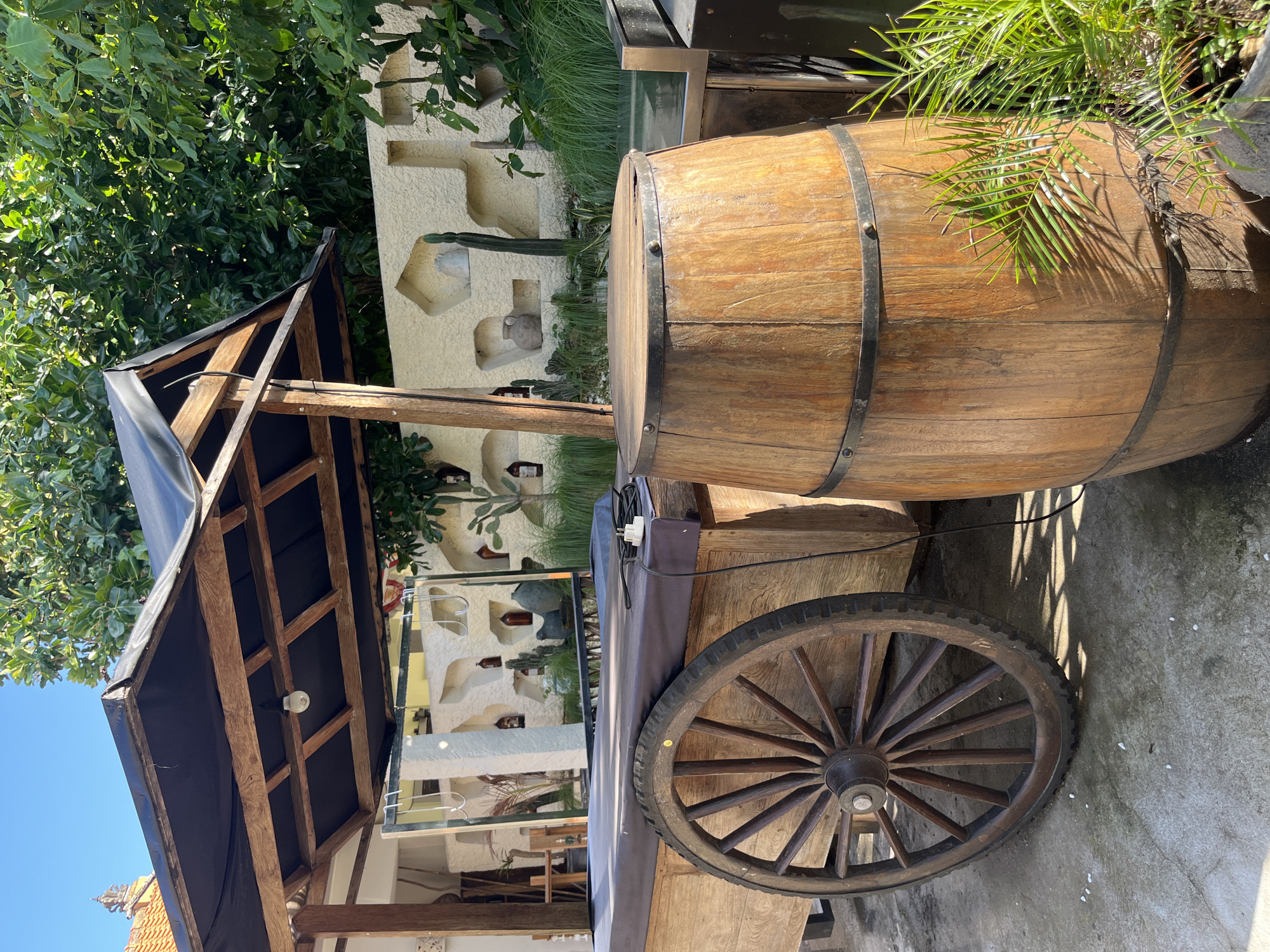 In Kuta Selatan, Indonesia, a wooden cart with a large spoked wheel is parked outdoors. The cart has a dark fabric covering and a wooden frame supporting a black roof. Adjacent to the cart is a wooden barrel with metal bands. In the background, a textured wall features decorative niches and potted plants, including a cactus. Lush green trees and a clear blue sky complete the outdoor scene.