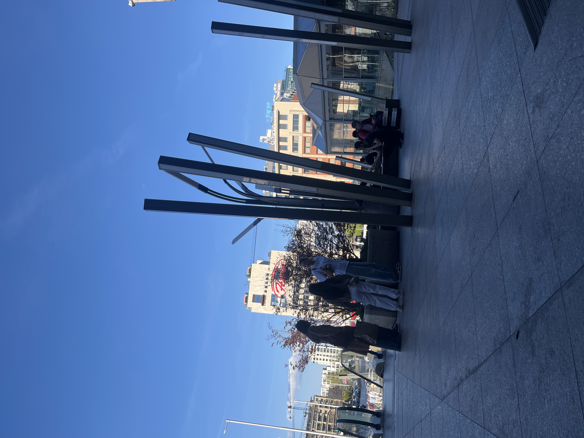 The image shows an outdoor scene in Sofia, Bulgaria, featuring a modern urban environment.
**Location:** The setting appears to be a public plaza or walkway within the city, identifiable by the contemporary architecture and urban infrastructure.
**People:** There are several people present in the image, but they appear in the background.
**Subjects:** The primary subjects include:
* Modern metal sculptures or structures framing the view.
* Buildings in the background, some with recognizable logos or signage.
**Objects:** Noteworthy objects include:
* Large commercial buildings.
* Metal structures/art installations.
**Actions:** People seem to be walking.
**Interactions:** No distinct interactions can be determined from this image.
**Environment:**
* **Weather:** The weather seems clear and sunny, indicated by the blue sky.
* **Architectural style:** Modern architecture dominates the buildings.
* **Infrastructure:** Cranes are visible.
* **Vegetation:** There are trees with branches in the image.