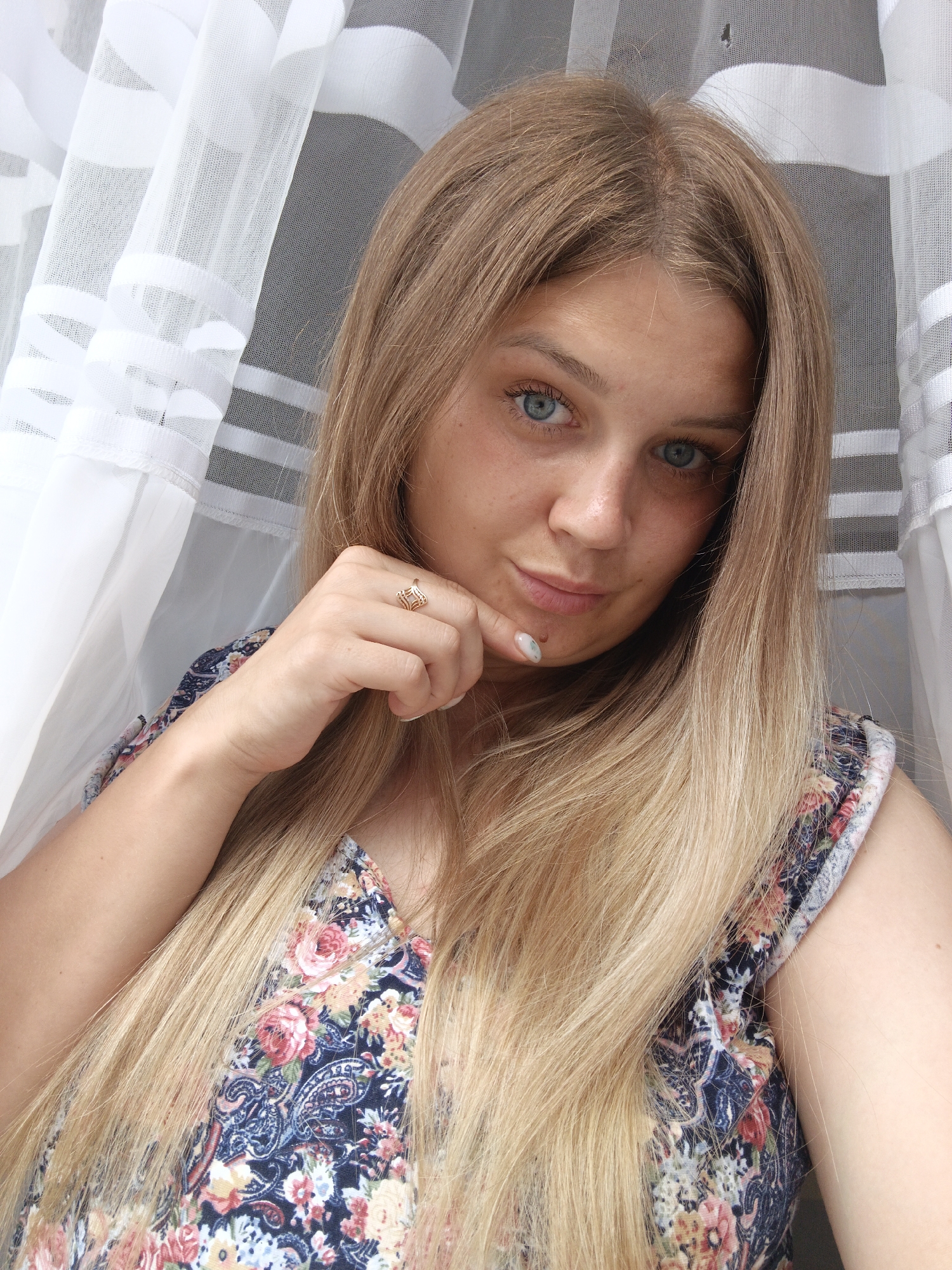 A young Caucasian woman with long blonde hair, blue eyes, and fair skin is shown in a close-up portrait. She is wearing a floral print dress with blue, red, and yellow flowers. Her right hand is raised to her chin, and she is wearing a delicate gold ring on her ring finger. She has light makeup on, including mascara and lip gloss. Her fingernails are light-colored with a subtle pattern. The background consists of white sheer curtains with a striped pattern. The lighting appears to be natural. The setting is indoors, possibly near a window. There is no information about the location in Medvedyovka, Russia.