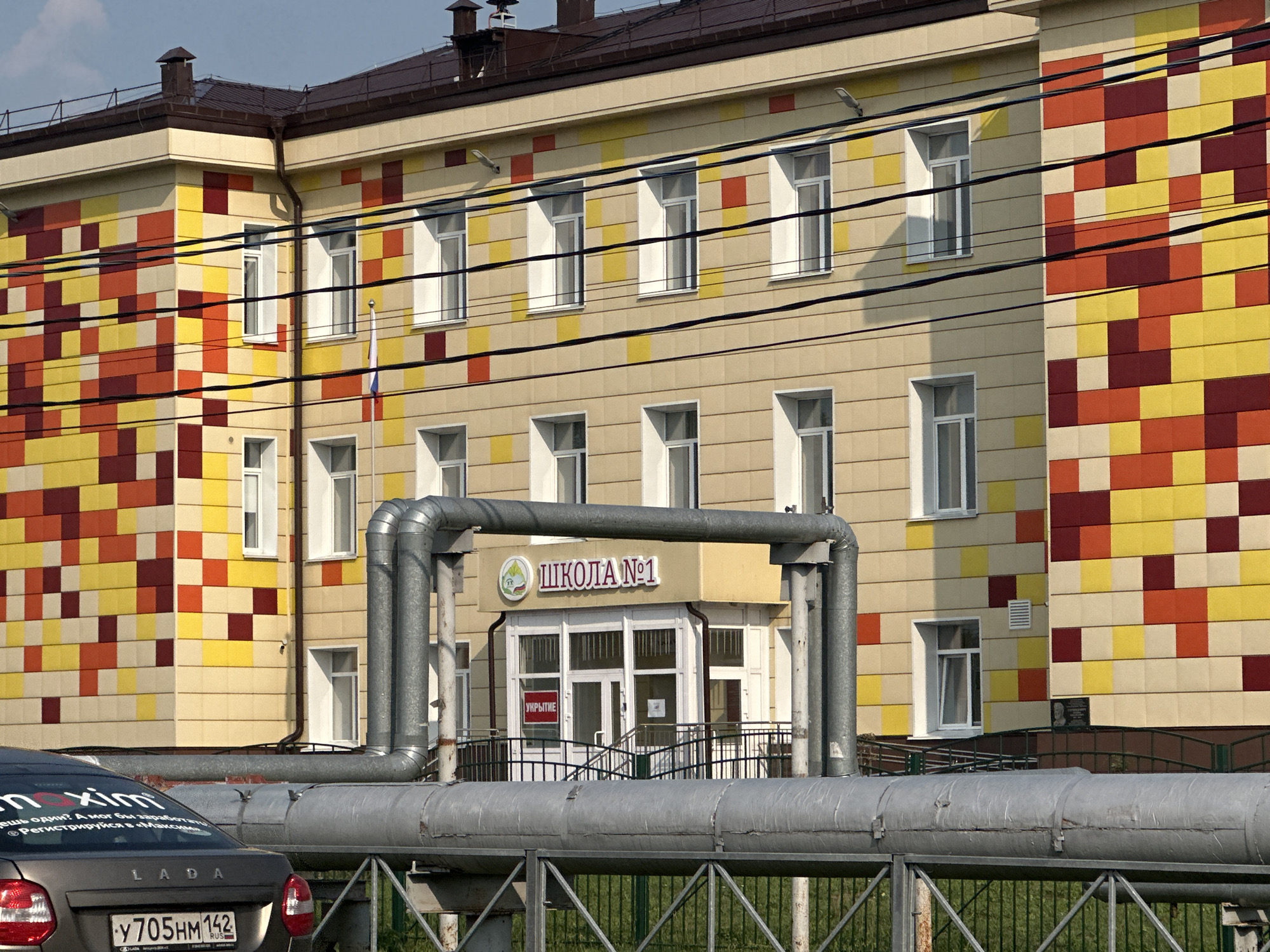 The image shows the exterior of a school building in Beryozovsky, Russia. The building has a colorful facade with a pattern of yellow, orange, and burgundy tiles. There are several windows on the building, and a sign above the entrance reads "ШКОЛА №1" (School No. 1). In the foreground, there is a gray car with a license plate that reads "Y705HM 142". Large metal pipes are visible in front of the building, and a green fence is in the foreground. The sky is visible at the top of the image, and there are some trees and grass in the background.