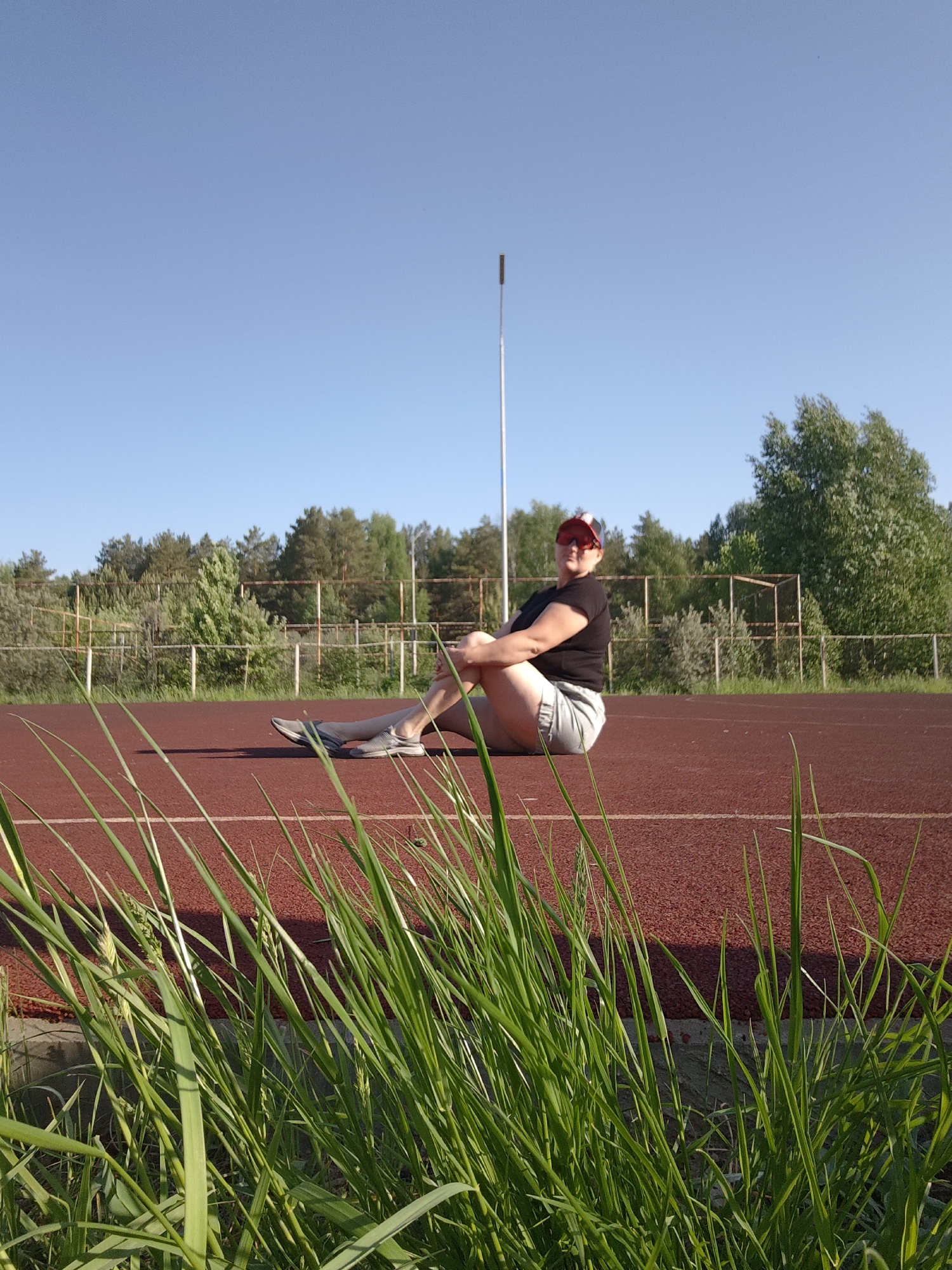 The image shows a person sitting on a running track in Markovskii, Russia. The person is wearing a black t-shirt, light-colored shorts, a red and white baseball cap, and gray sneakers. They are sitting with their legs crossed and arms around their legs. The running track is red with a white line. In the foreground, there is tall green grass. In the background, there are trees, a metal fence, and a metal pole. The sky is blue.