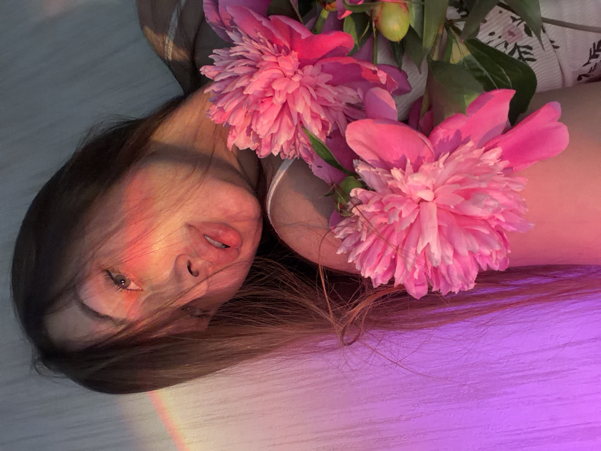 In the provided image, a young woman with long, brown hair is captured in a close-up shot. She is positioned at an angle, with her head tilted back and her face partially obscured by her hair. A pinkish-orange light casts a glow on the left side of her face, highlighting her eye and cheekbone, and a faint rainbow effect is visible. Her lips are slightly parted, revealing a hint of her teeth. She is holding a bouquet of vibrant pink peonies, with their delicate petals and green leaves filling the foreground. The woman is wearing a white top with a floral pattern. The background appears to be a textured wall, illuminated with a soft purple light. There is no identifiable information within the image to indicate its specific location in Chaikovsky, Russia.