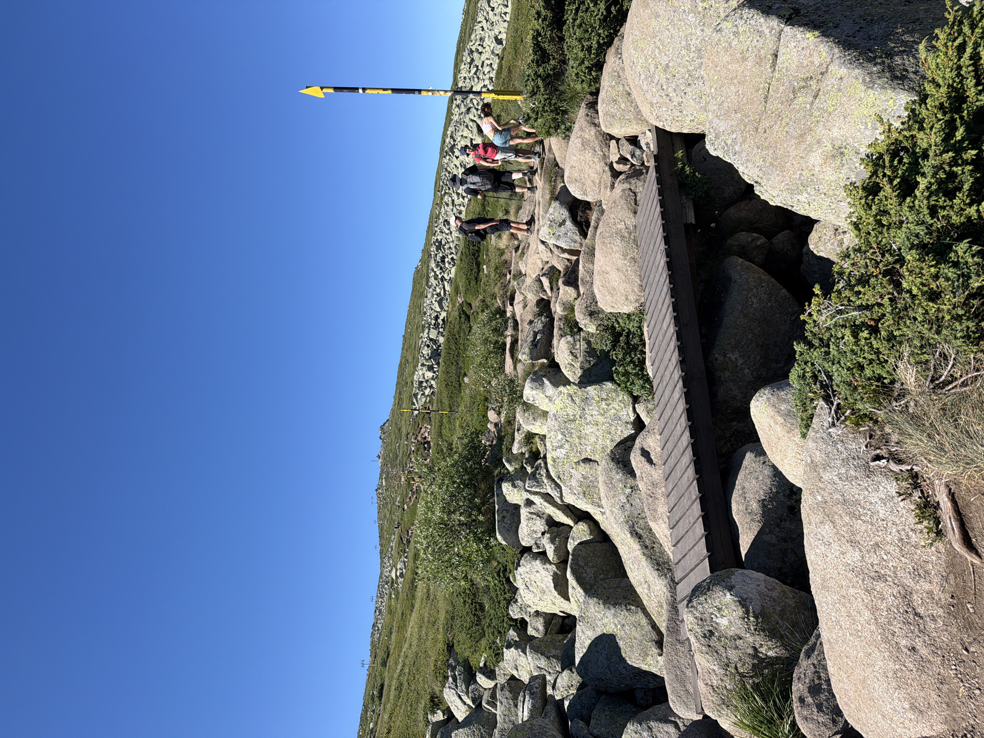 The image depicts a mountainous landscape with a clear blue sky. In the foreground, a wooden footbridge crosses over a rocky area with large boulders. To the right of the bridge, a yellow and black striped pole with a yellow triangle at the top stands vertically. Further in the background, a group of four hikers are visible, walking on a rocky path. The path ascends a grassy slope dotted with more rocks and sparse vegetation. In the far distance, the silhouette of a mountain peak is visible. The overall scene suggests a hiking trail in a high-altitude environment, likely part of the Vitosha mountain range near Bistritsa, Bulgaria. The hikers are dressed in outdoor clothing and appear to be engaged in a hike.
