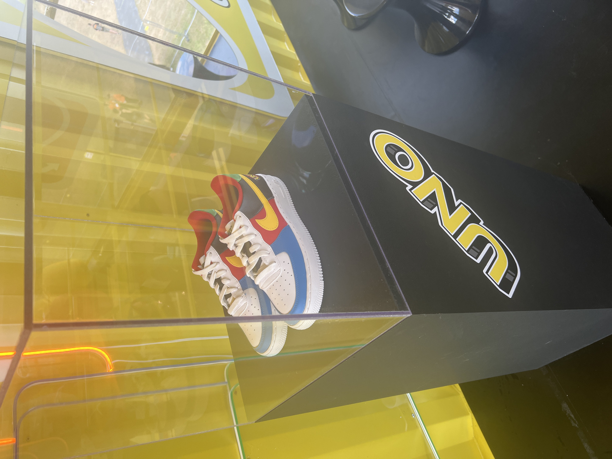 In Gdynia, Poland, a pair of colorful Nike Air Force 1 sneakers are displayed in a clear acrylic case. The sneakers feature a white base with red, blue, yellow, and black color blocking, and yellow Nike swooshes. They sit on a black pedestal with the "UNO" logo in yellow and white. The background is a vibrant yellow, with an orange neon light visible on the left. Reflections of people and outdoor scenery are visible on the case's surfaces, suggesting the display is in a public area.