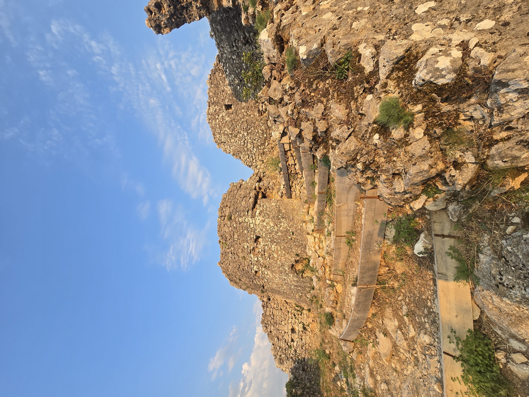 The image captures a section of the Medun fortress ruins in Medun, Montenegro, under a clear blue sky with scattered white clouds. The foreground is dominated by rugged, rocky terrain and the weathered stone walls of the ancient fortress. Wooden planks, possibly part of a restored pathway or protective barrier, are visible, forming curved sections that ascend the rocky slope. These planks are reinforced with metal rods. Small patches of green vegetation are growing amongst the rocks and ruins. The stone structures themselves are composed of rough-hewn stones, some appearing to be crumbling or partially collapsed, testament to their age. The warm, golden light suggests it is either sunrise or sunset, casting long shadows and highlighting the texture of the stones. The overall scene evokes a sense of historical significance and rugged natural beauty.