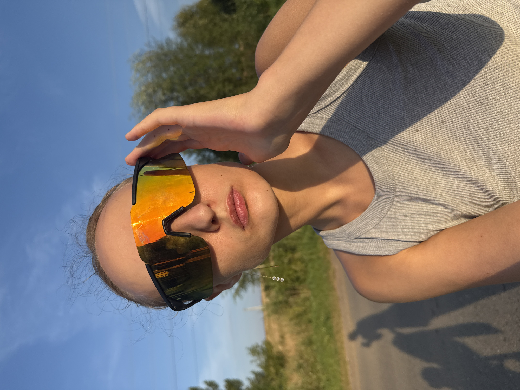 The media file shows a close-up, rotated image of a person wearing mirrored sunglasses and a grey t-shirt. The person's right hand is raised, adjusting the sunglasses, with their fingers partially covering the lenses. The sunglasses are large and wrap around the face, with orange and yellow reflections. The person's lips are slightly parted. A delicate earring with three small, clear stones hangs from their earlobe. The background suggests an outdoor setting with a bright blue sky and some greenery, possibly trees and fields, indicating a daytime scene. A paved road is visible in the lower portion of the image, and a shadow of what appears to be two figures, one with outstretched arms, is cast on the road. The location is identified as Podshivalovo, Russia. The overall impression is casual and summery, with the sunlight and outdoor environment suggesting pleasant weather. There is no visible text in the image.