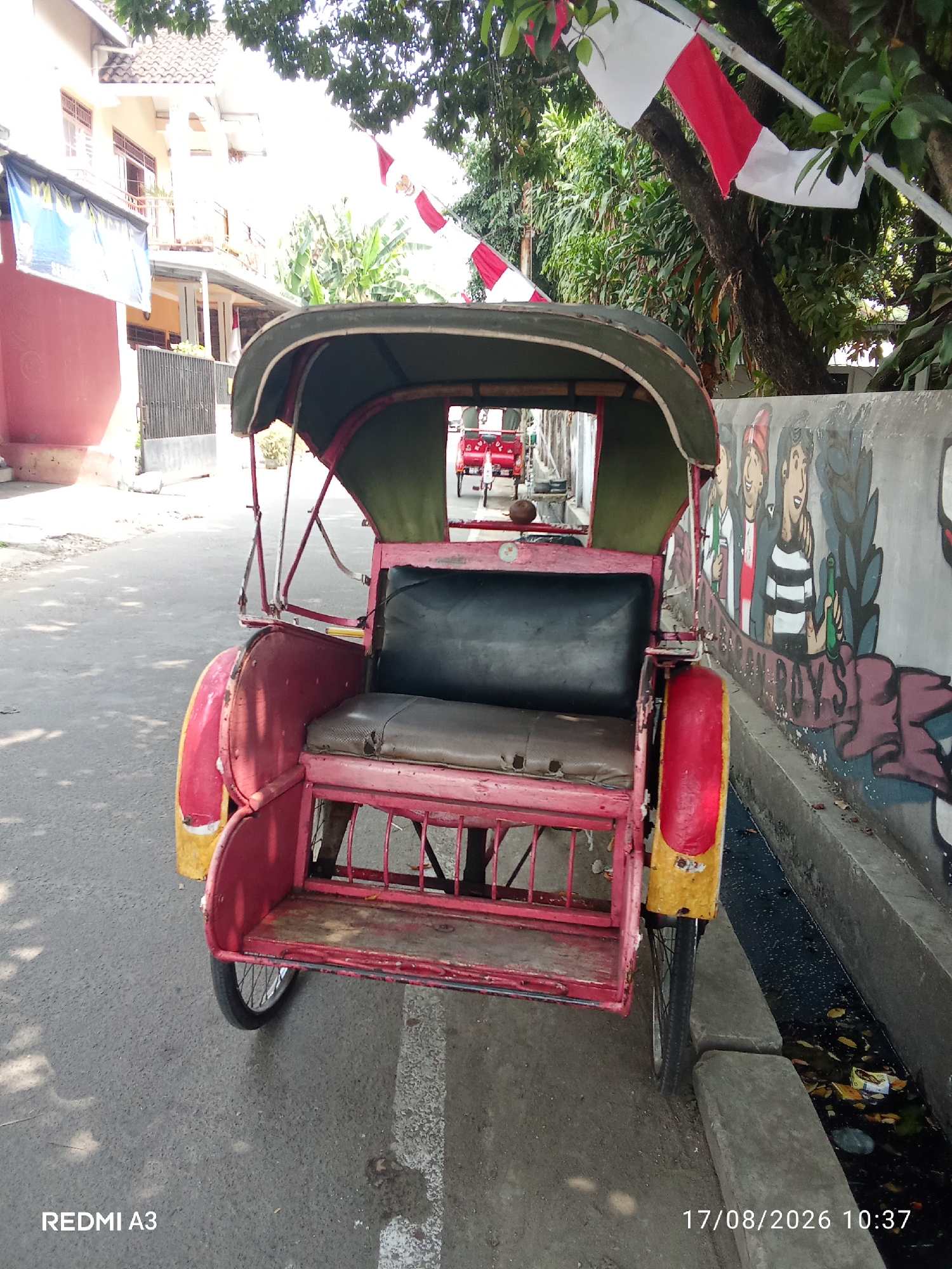 An outdoor, eye-level, medium shot shows a red and yellow tricycle parked on an asphalt road. The tricycle has a green canopy, a black seat, and a wooden passenger bench. A chain-link fence and a pink building are visible in the background. A mural depicting cartoon characters is painted on a wall to the right of the tricycle. Indonesian flags are hung from a tree above the tricycle. The image is timestamped 17/08/2026 10:37 and branded "REDMI A3". The scene is located in Banaran, Indonesia.
