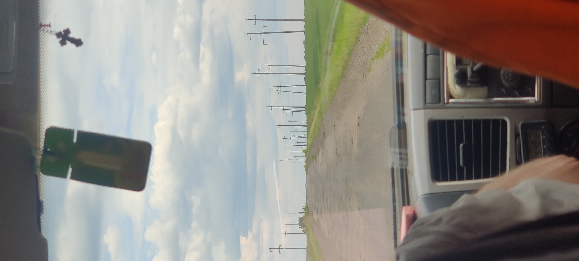 The image captures a sideways view from inside a vehicle, looking out onto a rural landscape in Shelestove, Ukraine. The scene is dominated by a long dirt or gravel road stretching into the distance, bordered on one side by a lush green field, likely agricultural. A continuous line of wooden utility poles with overhead power lines runs alongside the road, extending towards the horizon under a bright sky with scattered white clouds. A faint white contrail is visible in the sky.
Inside the vehicle, elements of the interior are visible. In the upper left, a dark, ornate cross hangs from a chain, possibly from the rearview mirror. Below it, a blurry, rectangular object with a green tint, possibly a phone or a pass, is seen. On the right side, parts of the vehicle's dashboard are visible, including a silver-colored air vent with vertical slats and a small, unreadable digital display unit. An orange piece of fabric or trim is in the upper right corner. In the lower right, a person's forearm and hand rest inside the vehicle, with a white, soft object (like a cloth or tissue) nearby.
The image portrays a daytime scene, suggesting fair weather conditions during a journey or travel through the countryside. There is no discernible legible text within the frame.