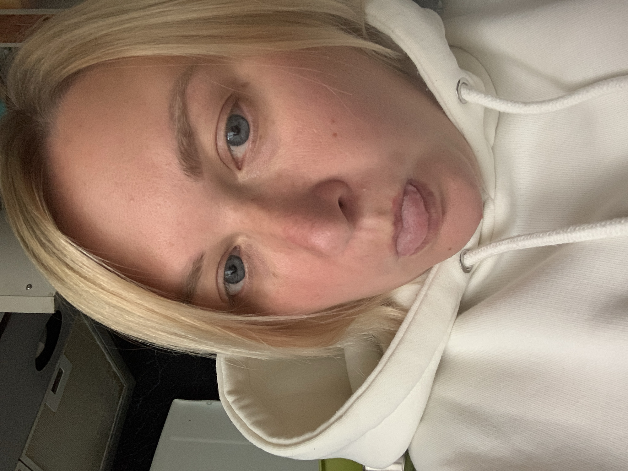 A selfie captures a fair-skinned individual with blonde hair and blue eyes, wearing a white hooded sweatshirt. Their tongue is slightly extended in a playful expression. The background reveals a glimpse of an interior space, including what appears to be a storage cabinet or drawer unit. The exact location in the city of Chaikovsky, Russia, is impossible to determine from the provided image.
