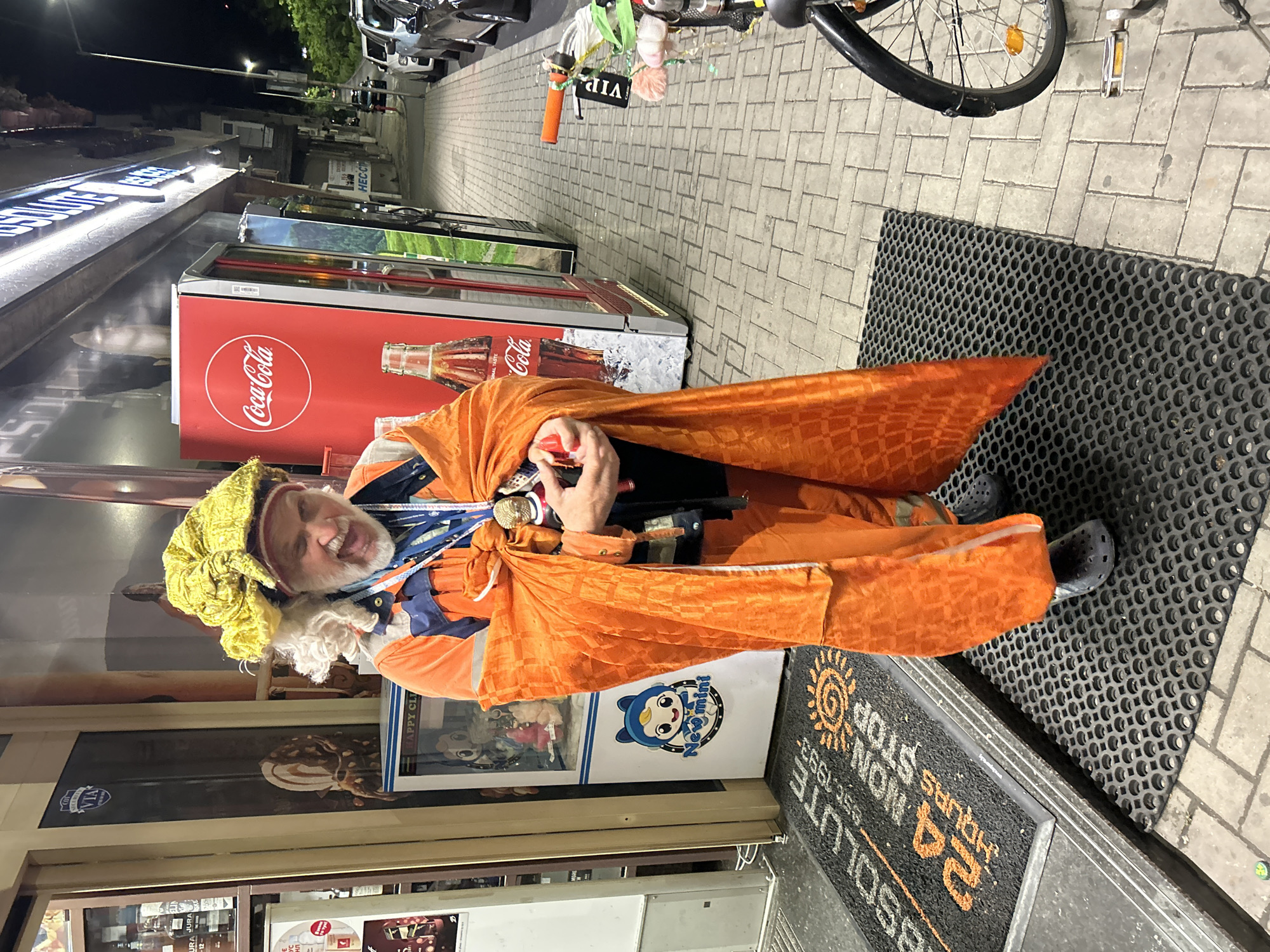 A man in an orange costume stands on a doormat that reads "Absolute" on a sidewalk. Behind him is a storefront with Coca-Cola refrigerators and advertising. The man wears a yellow head covering and orange cape. A bicycle is parked on the sidewalk, and cars are parked on the street in the background. The location appears to be an urban area at night.