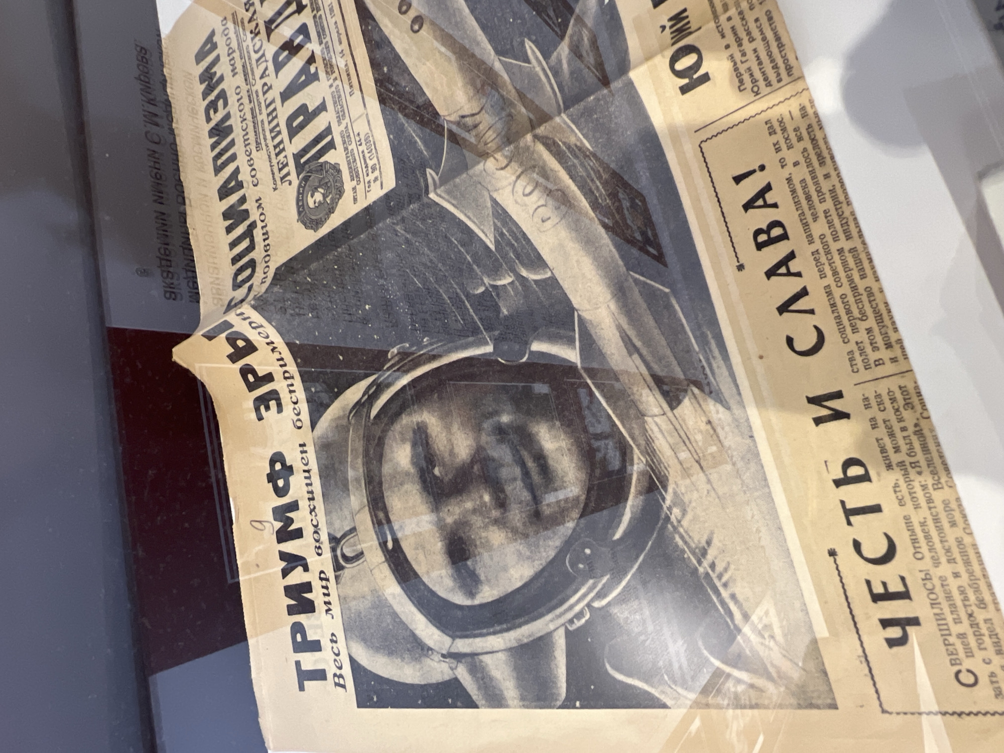 This is a photograph of a preserved page of the Soviet newspaper "Pravda". The newspaper page features a headline reading "TRIUMPH OF ERA" and displays a portrait of a cosmonaut, possibly Yuri Gagarin, within a spacesuit helmet. An image of a rocket is superimposed over the portrait. The other side of the page includes text that celebrates the achievements of Soviet cosmonautics with the words "HONOR AND GLORY!". The newspaper is probably an exhibit in a museum located in Saint Petersburg, Russia, that covers Soviet history.