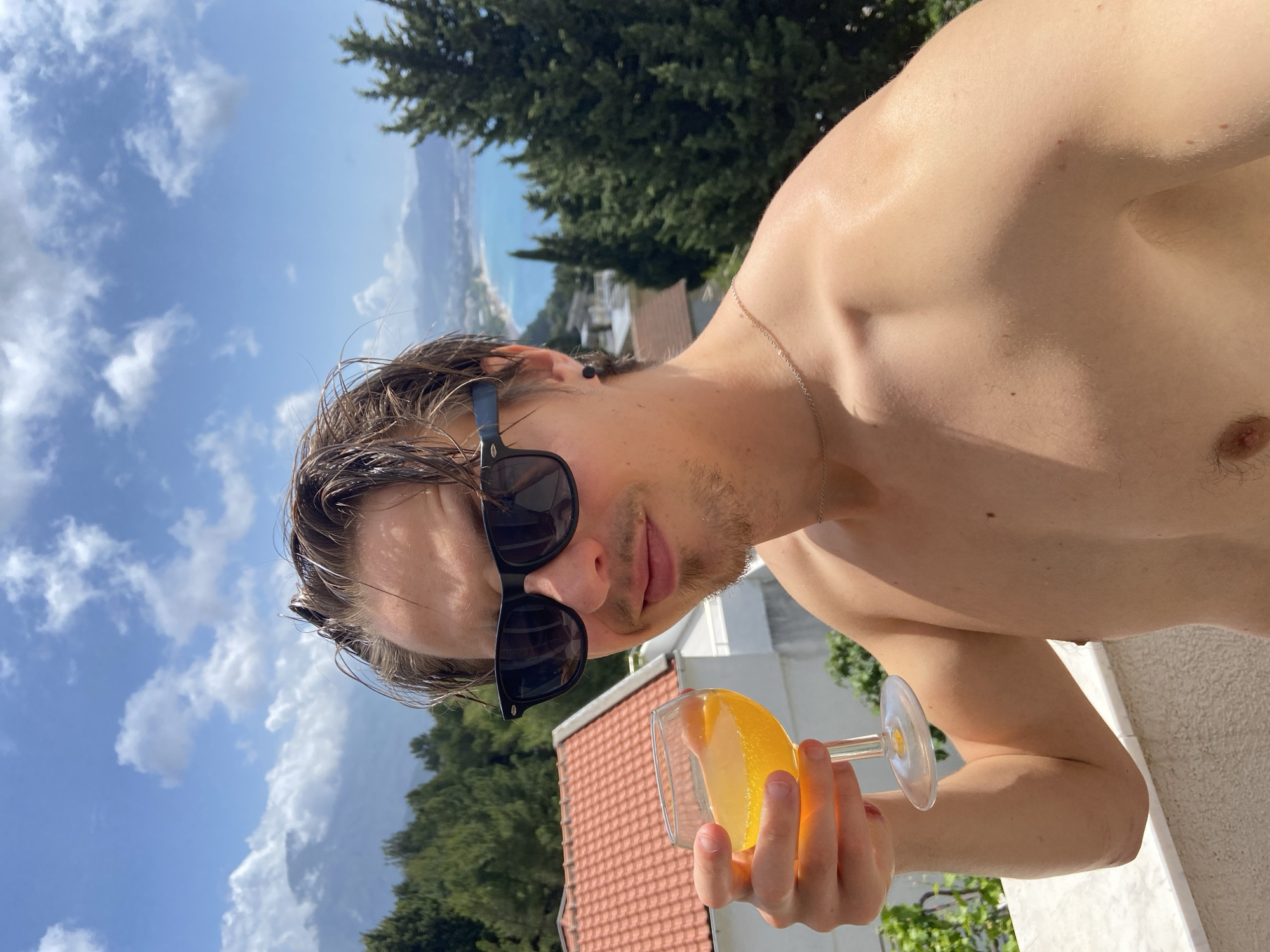 A shirtless man wearing sunglasses is holding a glass of orange juice. He appears to be on a balcony overlooking a view of buildings, trees, and the coastline. Based on the visual cues, the location is likely Šušanj, Montenegro. The sky is partly cloudy.