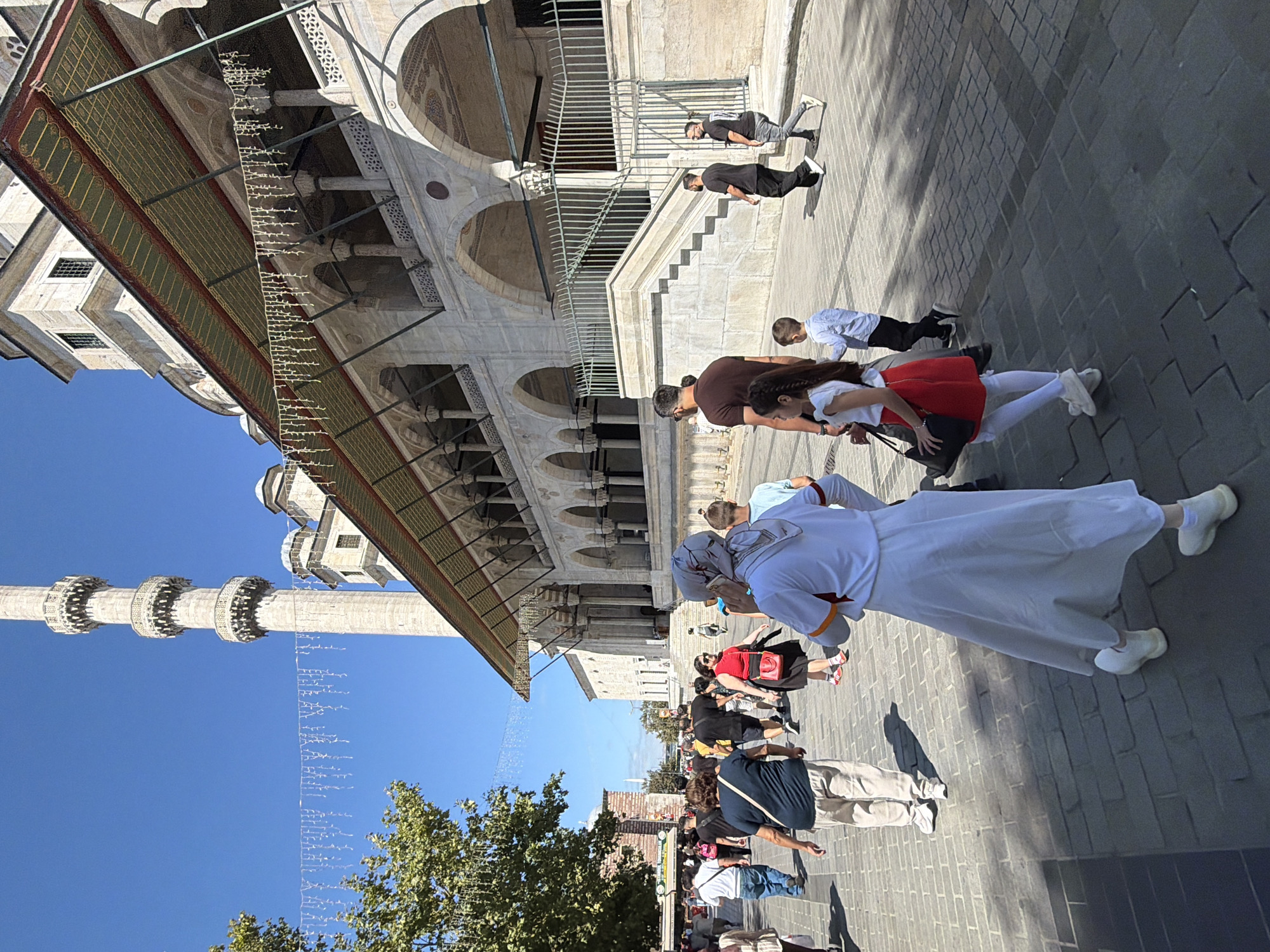 The image depicts a scene in front of the Suleymaniye Mosque in Istanbul, Turkey, under a clear blue sky. A minaret of the mosque is visible on the left, with decorative strings of lights hanging from the building's overhang in the foreground. The mosque's architecture, characterized by arches and intricate stonework, dominates the right side of the frame. A paved walkway, made of grey cobblestones, is populated by a diverse group of people. In the foreground, a woman wearing a white abaya and a headscarf is walking away from the camera, her face partially obscured. Beside her, a young girl in a red skirt and white top walks with a black handbag. Several other individuals, including men, women, and children, are dispersed throughout the walkway, some walking towards the mosque, others walking away. Some individuals appear to be tourists, evident from their casual attire and the presence of a camera bag on one woman. The atmosphere appears to be one of a bustling public space, likely a tourist attraction, with people engaging in various activities such as walking, talking, and observing the surroundings. The sunlight casts distinct shadows on the cobblestone pavement, indicating a bright, sunny day.
