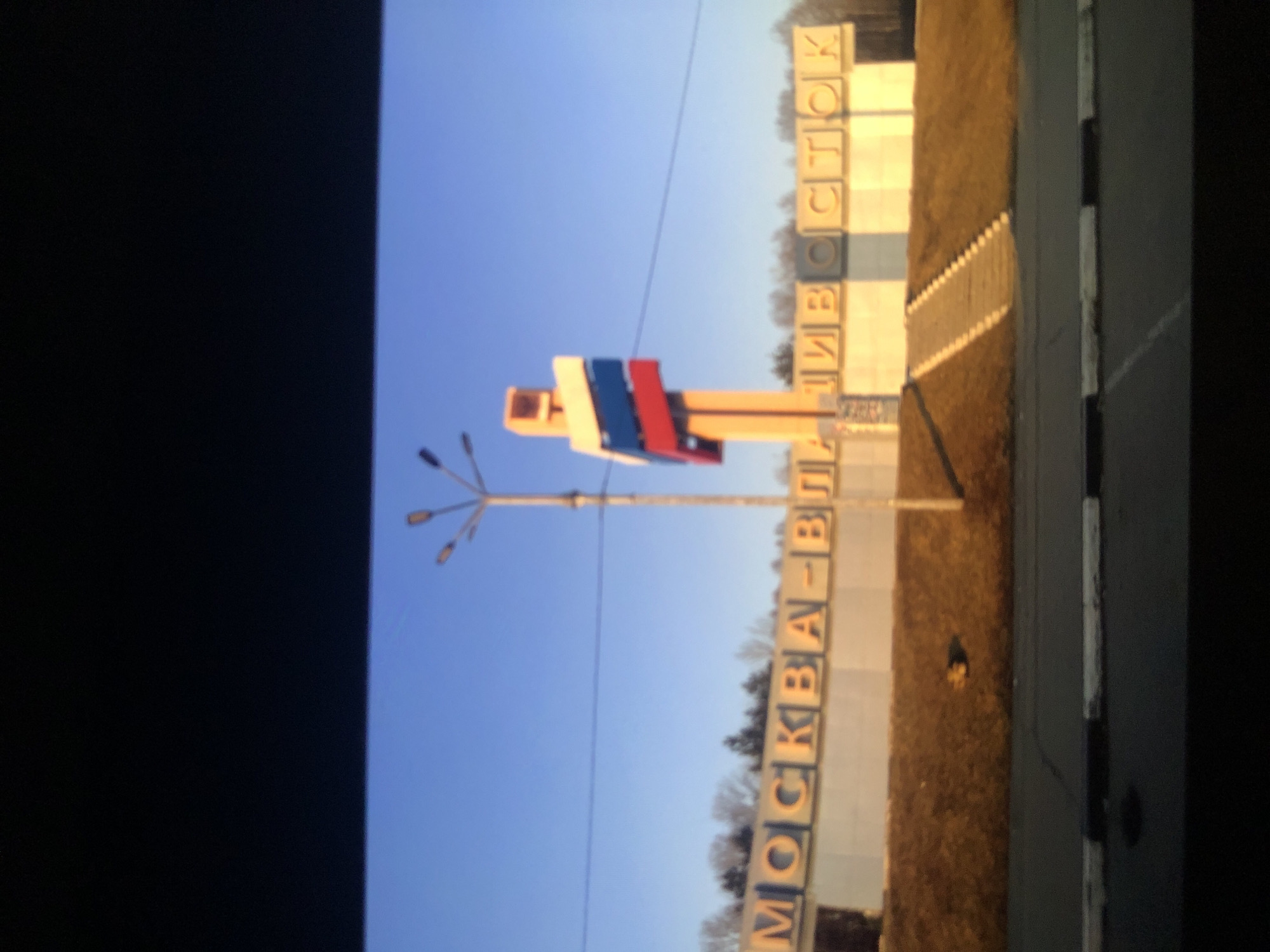A roadside sign marks the entrance to Vladivostok from Moscow, Russia. The sign is in Russian. A street lamp is positioned in front of the sign. The sky is blue. A curb or road edge borders the sign.