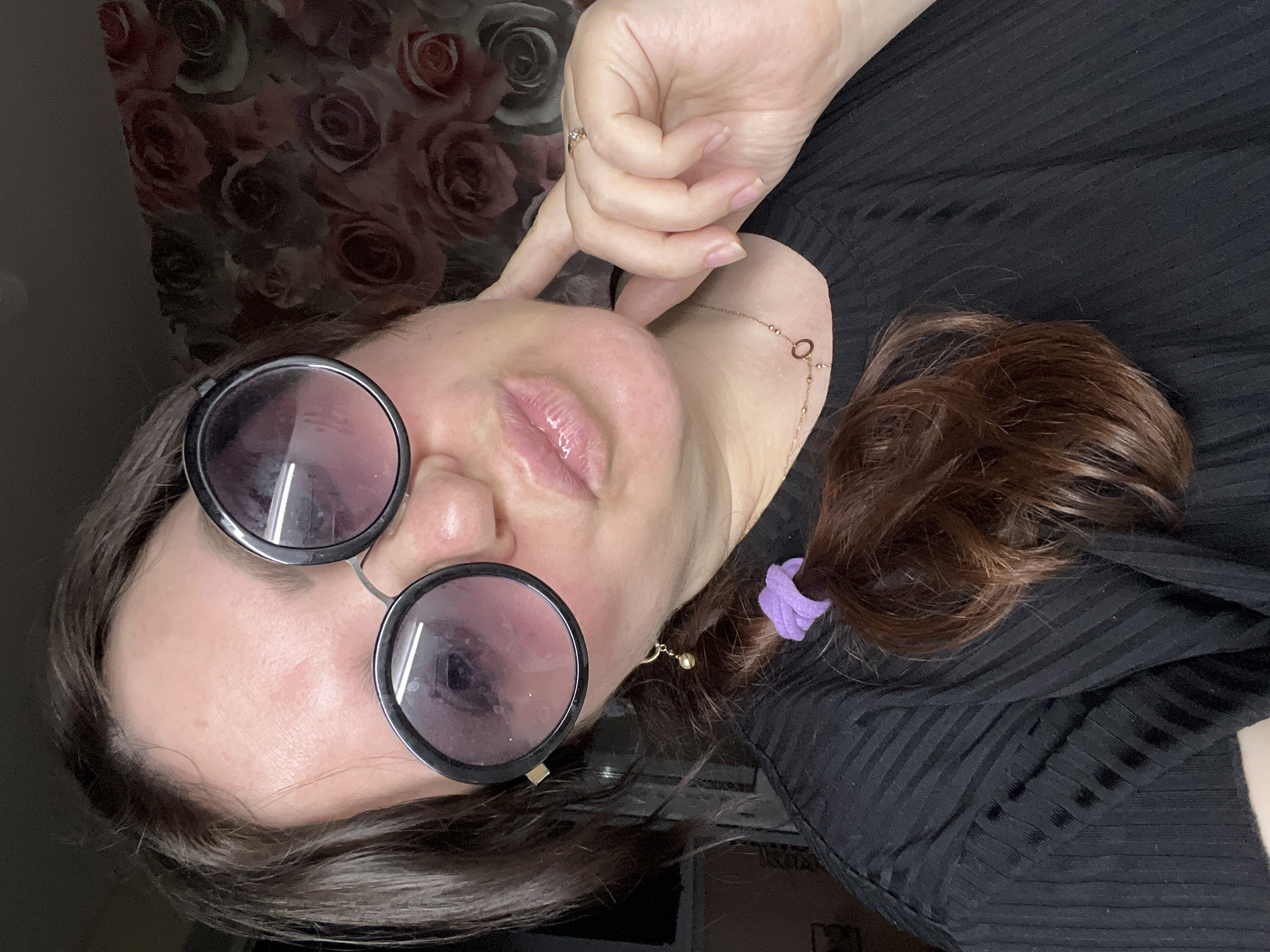 A woman with shoulder-length brown hair, partially tied back with a purple scrunchie, poses close to the camera while wearing round, dark-rimmed glasses with visible smudges on the lenses. She wears a black, ribbed shirt and a delicate gold chain around her neck, with a ring on her finger. One hand is near her face, and she is positioned against a backdrop of a floral-patterned wall. No specific information indicates the city of Noviy, Russia, is associated with the image.