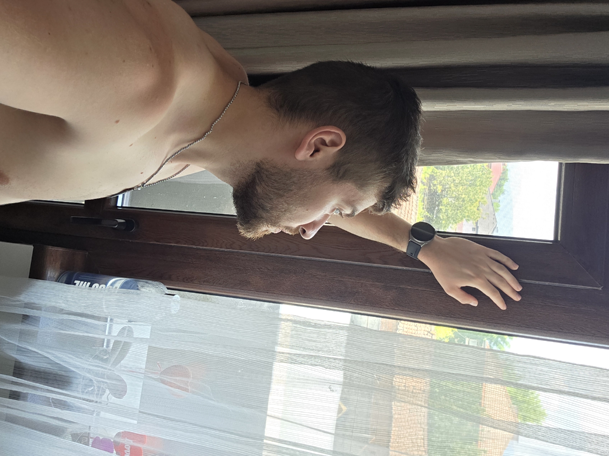 A male individual is observed in a right-side profile, standing bare-chested and looking out a window in Yagodovo, Bulgaria. He has short, dark brown hair and a matching beard. A silver chain necklace is visible around his neck. His left arm is raised, and his left hand, wearing a dark-colored smartwatch, rests against the dark brown window frame. To the left, a heavy, textured light brown curtain is drawn. To the right, a sheer white curtain partially obscures the view through the window. On the dark brown windowsill, behind the sheer curtain, a blue water bottle marked "1700 ML" and other indistinct objects are visible. The exterior view reveals a bright sky, green trees, and residential buildings with tiled roofs in the middle ground, suggesting a daytime, suburban setting.