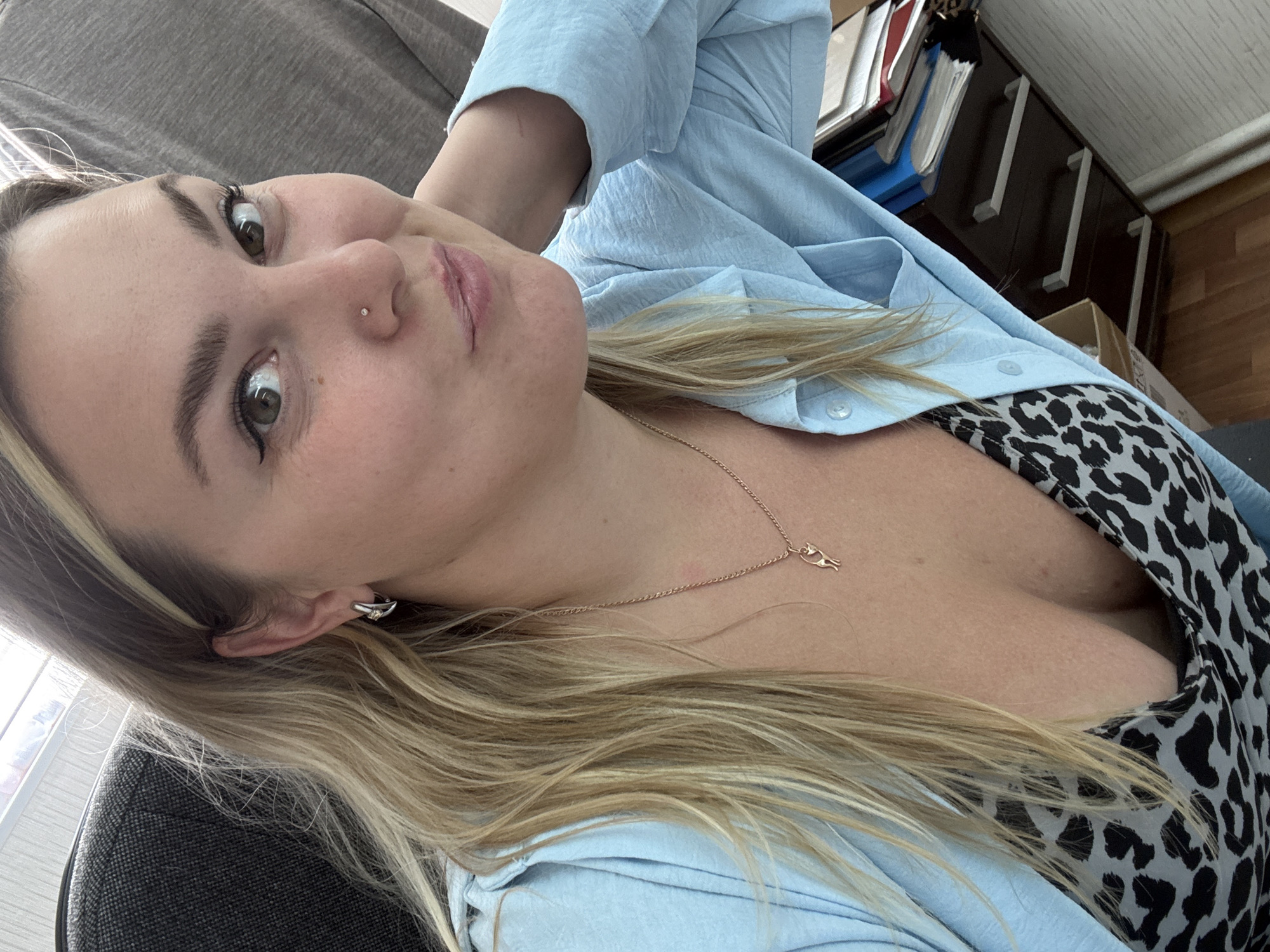 A woman with blonde hair is shown from the chest up, looking towards the camera. She is wearing a light blue collared shirt over a leopard-print top. A delicate necklace with a small cat-shaped pendant is visible. She has subtle eye makeup and a nose piercing. The background includes a dark office chair, a filing cabinet with papers, and a window. The image appears to be taken indoors, likely in an office or home setting, and does not provide any specific information about the city of Noviy, Russia.