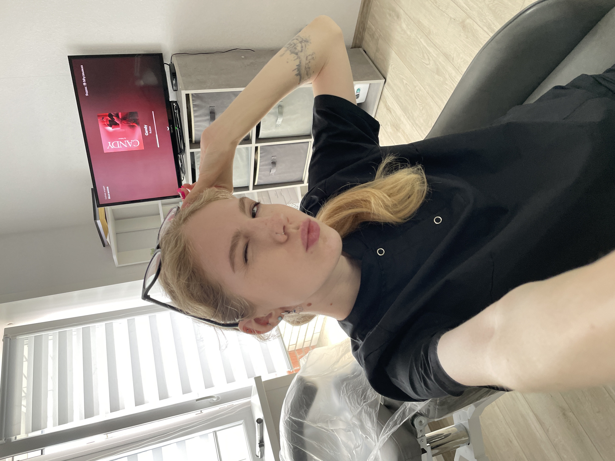 A blonde woman wearing black glasses and a black top is seated, positioned in the lower left of the frame. Her right arm is raised. She is looking and winking toward the camera, puckering her lips. A tattoo is visible on her raised arm.
Behind her is a window with vertical blinds. Further in the background, there is a television showing the cover of a song called "Candy" by Ruben, played on Я.Музыка. The text on the screen is in Russian. Next to the TV is a storage unit with drawers. The room has light-colored walls.
Based on the Russian text on the TV, it is likely that the location is within Russia. Given the specific service Я.Музыка (Yandex Music) is popular in Russia, the city could be anywhere, but assuming a larger population center where these services are common, it could be Izhevsk. However, this is inferential and cannot be confirmed definitively from the image.