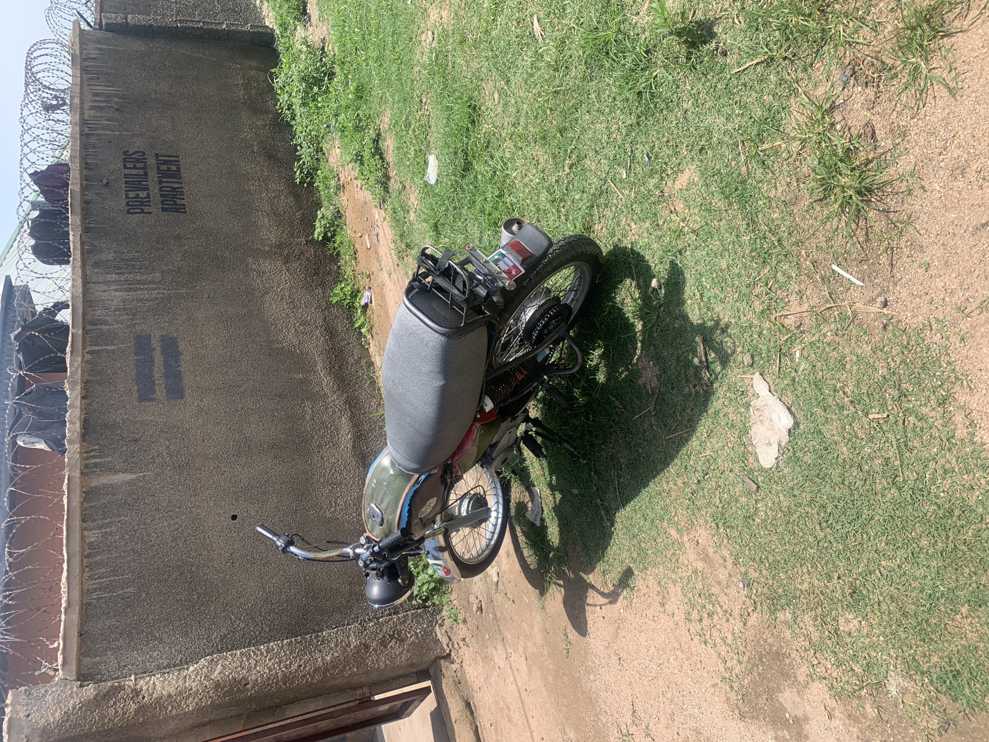 A motorcycle is parked on a patch of grass next to a dirt path. The motorcycle is a dark green color with black accents and a gray seat. It is facing towards the right side of the image. Behind the motorcycle is a weathered concrete wall with barbed wire on top. The wall has the words "PREVAILERS APARTMENT" painted on it in black. The environment appears to be an outdoor area with dry ground and sparse grass. The lighting suggests it is daytime, with shadows cast by the motorcycle and other unseen objects. The general setting is a somewhat unkempt urban or semi-urban environment.