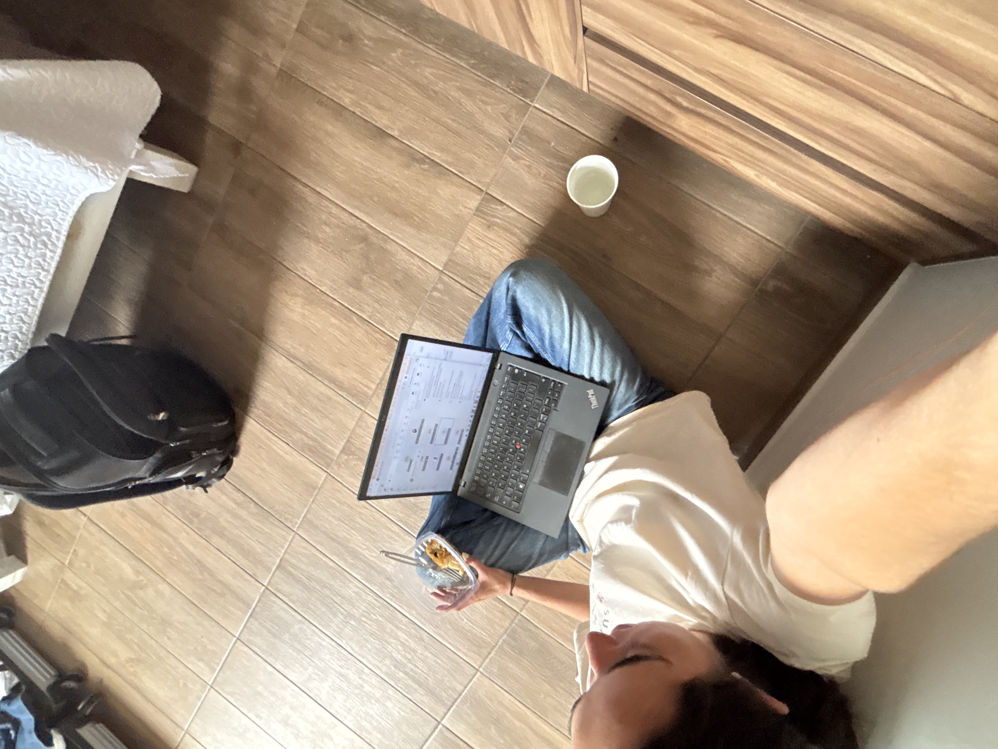 A high-angle, overhead shot depicts a person sitting on a wood-look tiled floor, seemingly working on a laptop. The individual is wearing blue jeans and a white t-shirt. A black ThinkPad laptop rests on their lap. They also hold a plastic container with food in their left hand. Beside the person, slightly to the left, is a black backpack. To the upper right is a white cup on the floor near what looks like a wooden cabinet. To the left is a white piece of furniture. No details in the image suggest the location is San Bernardino Tlaxcalancingo, Mexico.