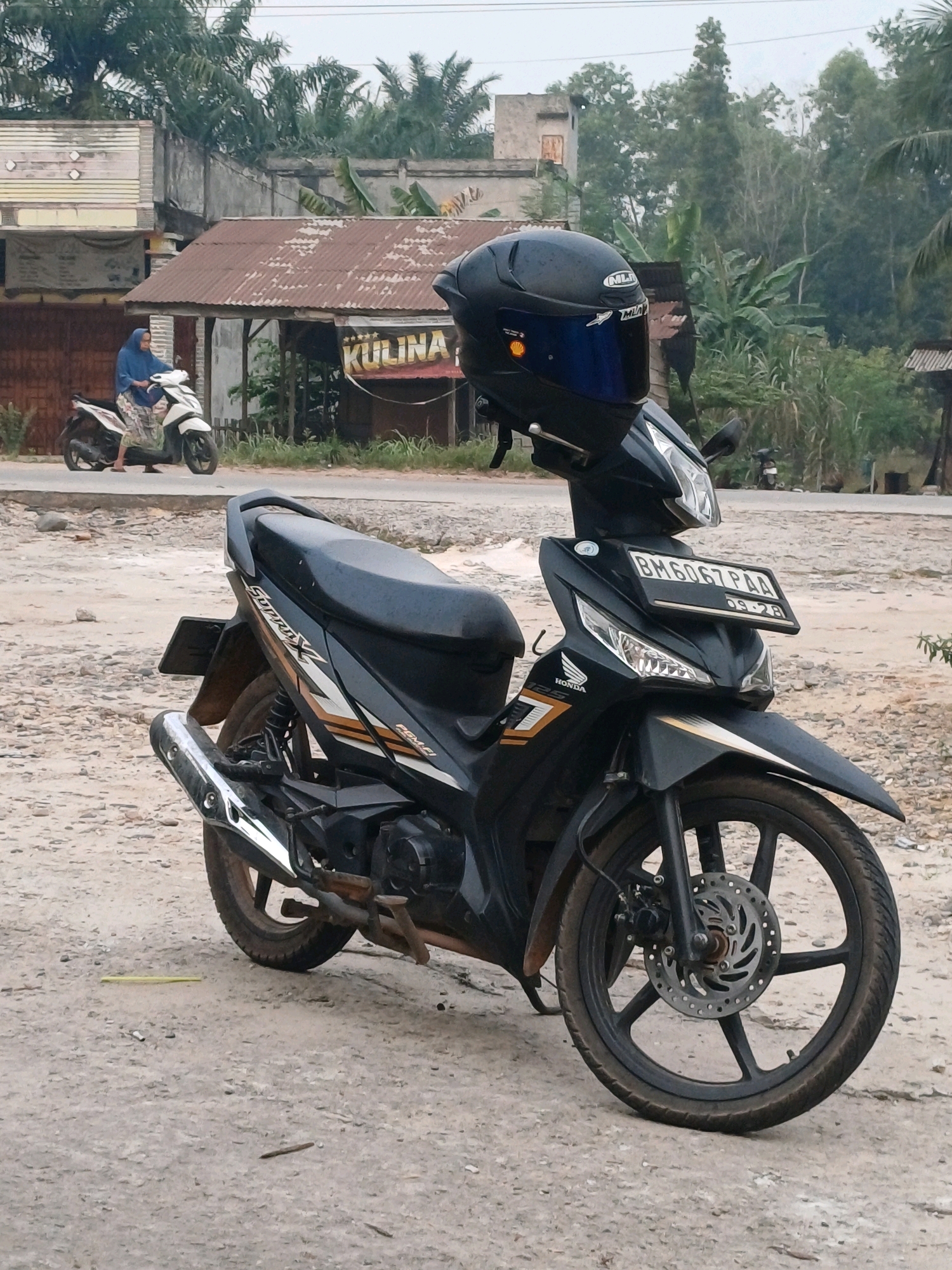 In Dumai City, Indonesia, an outdoor scene depicts a black Honda motorcycle parked on a dirt and gravel lot. A black helmet with a blue visor is placed on the handlebars. In the background, a woman wearing a blue hijab and patterned skirt sits on a white scooter. Behind her is a building with a rusty tin roof and a sign that reads "KULINA". Trees and other vegetation surround the area. The sky is overcast.