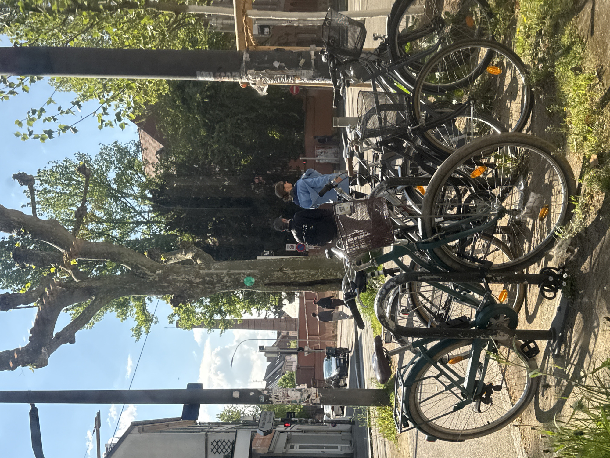 This daytime photograph captures a sunny urban street scene in Strasbourg, France. On the right side of the frame, numerous bicycles are neatly parked in a metal rack alongside a grassy strip. The bikes, predominantly dark-colored, feature handlebar baskets, rear racks, and orange reflectors on their wheels.
On the left, a thick, dark pole stands, covered with various stickers and some graffiti. A red circular "No Entry" sign is clearly visible on this pole, and below it, a white rectangular sign with illegible black text is affixed.
In the mid-ground, two individuals are walking away from the viewer on the sidewalk. One wears a light blue hoodie and sunglasses, while the other is dressed in a dark jacket and a baseball cap. A street extends into the distance behind them, with a dark-colored car visible further down. Buildings with brick facades and tiled roofs line the street, partially obscured by large, mature trees with lush green foliage. The sky is clear and blue with scattered white clouds, and the strong sunlight casts distinct shadows, suggesting either morning or late afternoon.
