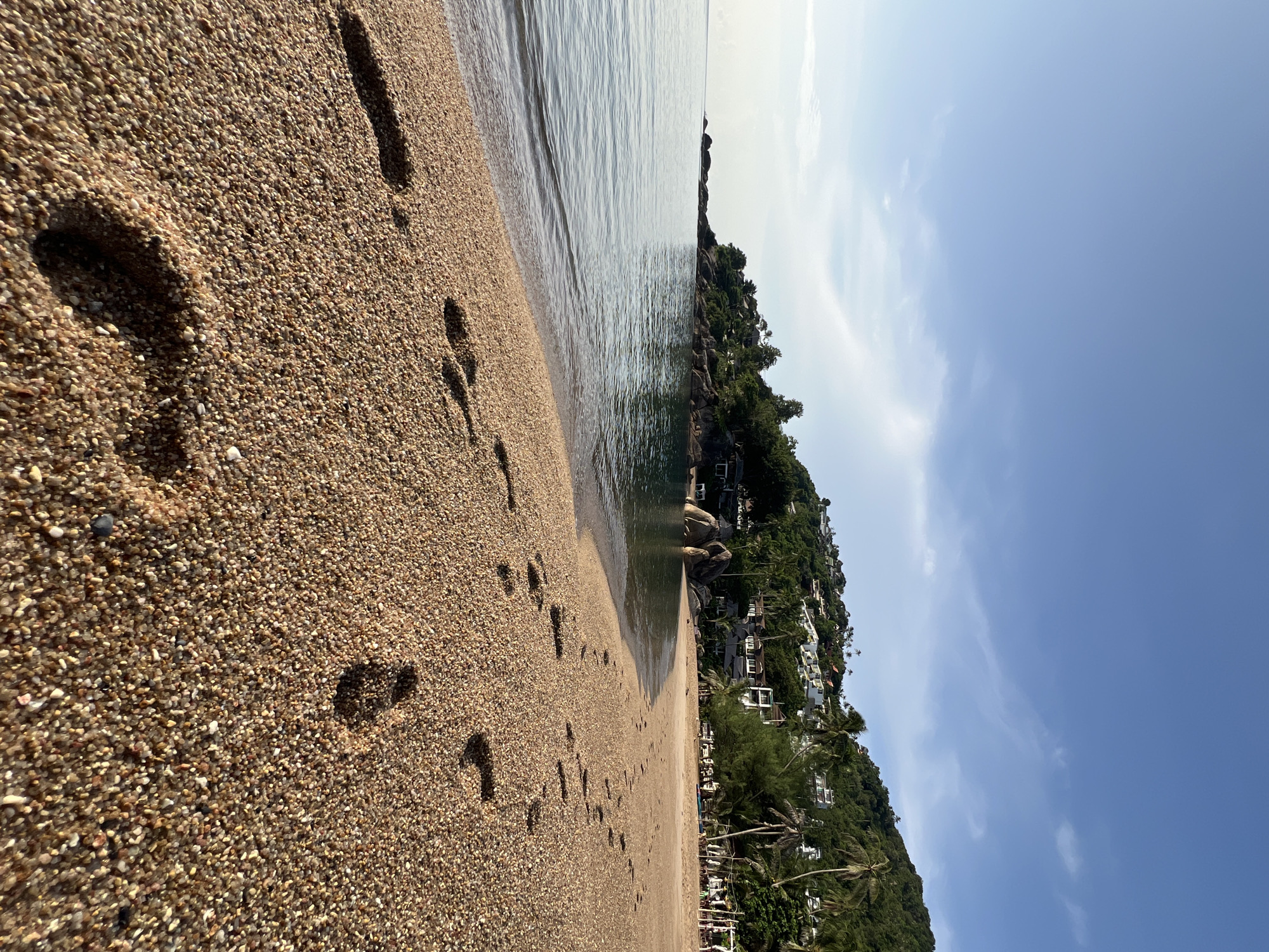 This is a vertical, eye-level, medium shot of a beach in the Ko Samui District of Thailand. The image is taken from the sand, looking out towards the ocean. In the foreground, footprints are visible in the light brown sand. The ocean occupies the left side of the frame and appears calm with small waves. Beyond the water, a rocky shoreline is visible, topped with lush green trees and houses scattered along a hillside. The sky is a clear blue with a few wispy white clouds. The overall scene depicts a serene tropical beach.
