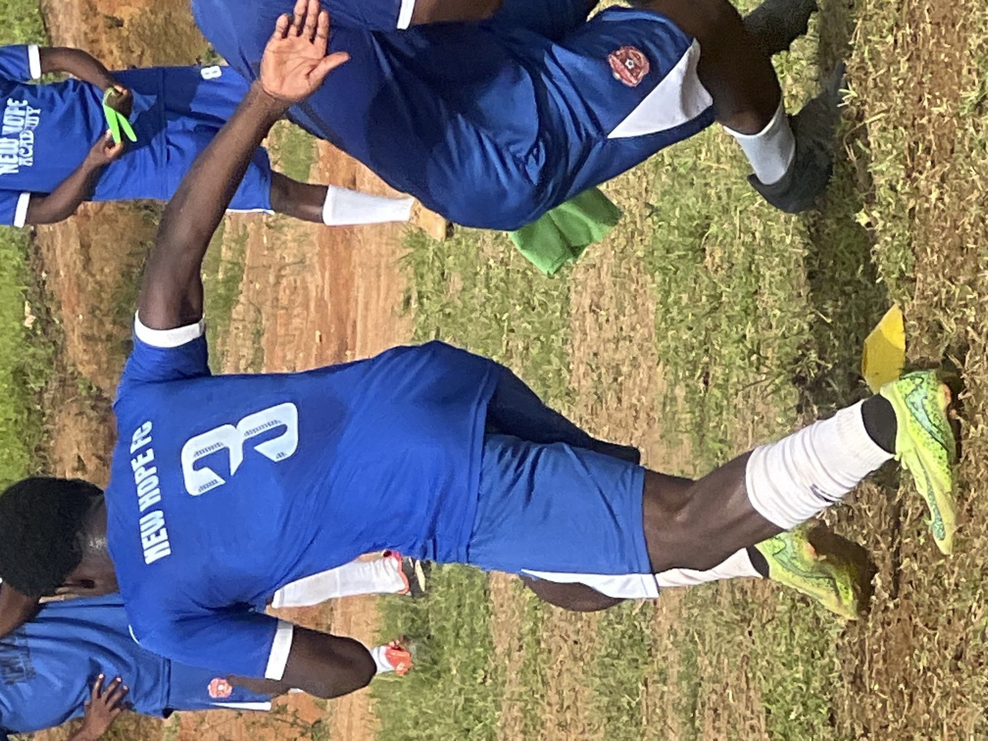 The image shows a close-up view of three male individuals in athletic attire, seemingly engaged in a soccer match or practice on a grassy field with dirt patches. The central figure, facing away from the camera, wears a royal blue soccer jersey with "NEW HOPE FC" emblazoned in white across the back, above the number "3." His shorts are also royal blue with white side panels. He is wearing white calf-high socks and vibrant lime green soccer cleats with a patterned design. His right arm is extended to his side, and his left arm is bent.
To his left, another individual is partially visible, also wearing a royal blue jersey with white trim. This person's right hand is visible, holding a neon green object, possibly a training marker.
To the right of the central figure, another person is seen from the waist down, wearing royal blue soccer shorts with a white panel and a red circular emblem with a soccer ball design. This individual is wearing dark soccer cleats and white socks. A folded green cloth is visible on the ground near their right leg.
The environment suggests an outdoor sports facility with dry, brown earth and scattered patches of green grass. The lighting indicates it is daytime. The overall scene depicts a moment during a sporting activity. There is no specific visual information in the image that allows for the determination of the location within the city of Jos, Nigeria.