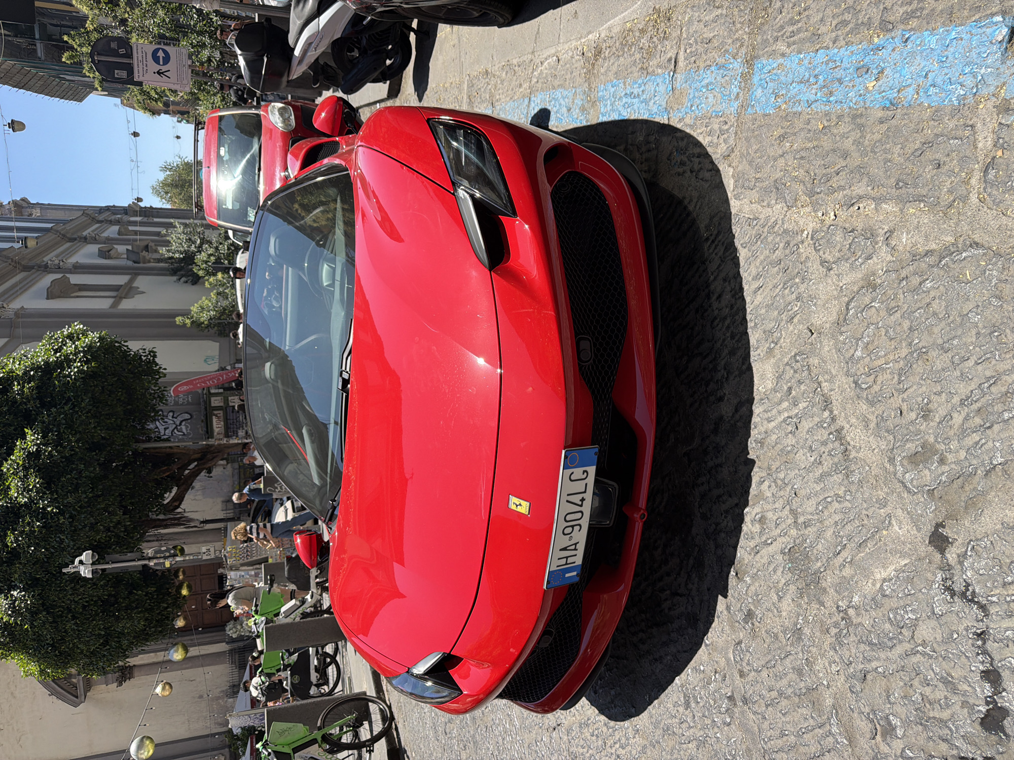 A red Ferrari sports car is parked on a street in Naples, Italy. The car is in the foreground, facing slightly to the left. The license plate reads "HA-904LG". Behind the Ferrari, a red van is visible, and further back, a white scooter is parked. The street is paved with cobblestones, and a blue painted parking space line is visible on the right side of the image. In the background, there are buildings, trees, streetlights, and several people walking or standing. The scene is illuminated by bright sunlight, casting shadows on the street.