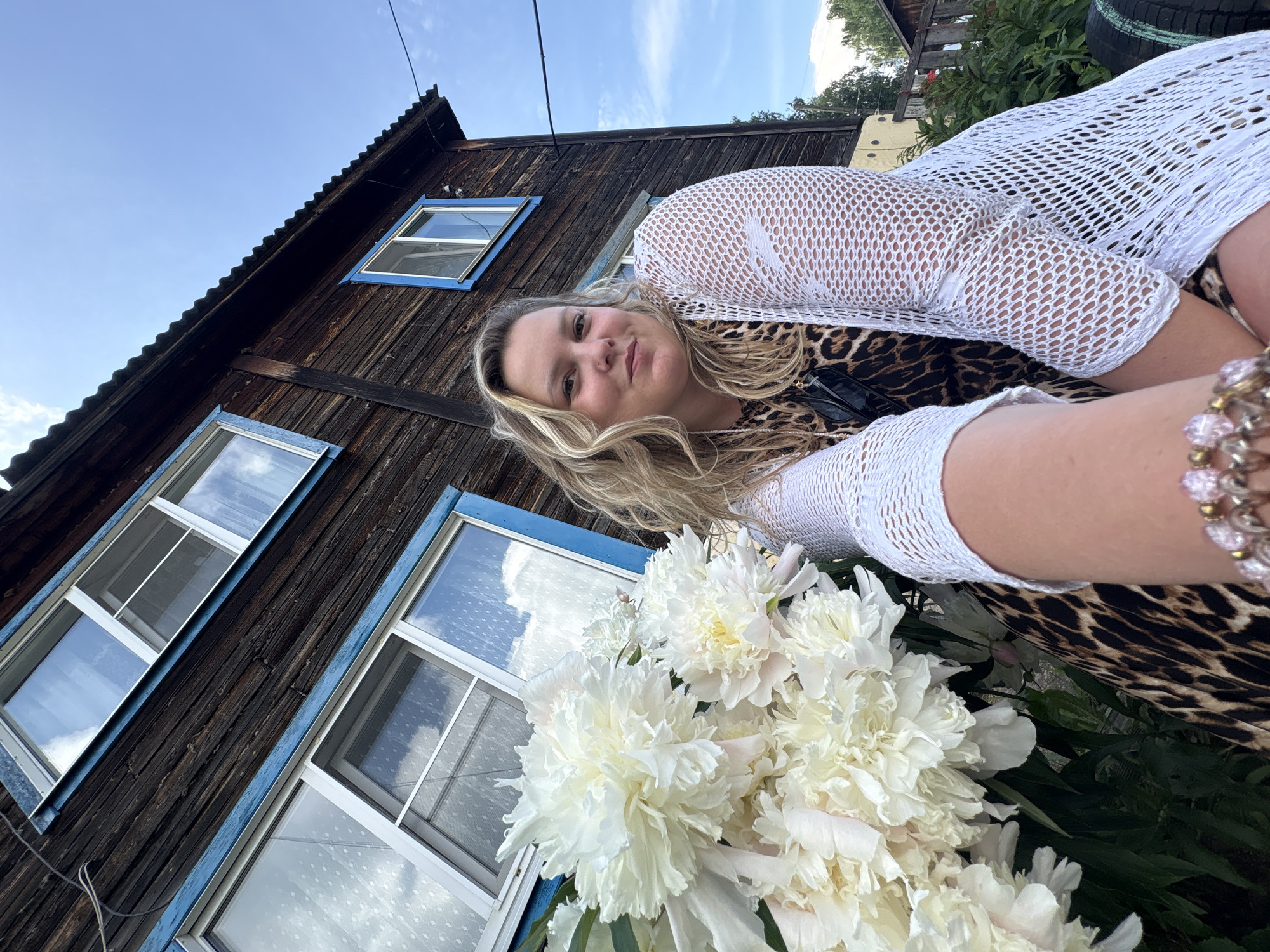 An outdoor, eye-level, medium shot shows a woman with blonde, wavy hair and fair skin posing in front of a weathered, dark wooden building with blue window frames and a corrugated metal roof. She is wearing a leopard-print dress and a white, open-knit cardigan. A large bouquet of white peonies is positioned to her left, obscuring part of the building. The background includes a clear blue sky with sparse white clouds. The setting appears to be a rural or suburban area, possibly a private residence. The wooden building suggests an older construction style. The woman is smiling directly at the camera, with her right arm extended towards the viewer as if taking a selfie. Her left hand rests on a bracelet adorned with pink beads. The scene conveys a casual, summery atmosphere.