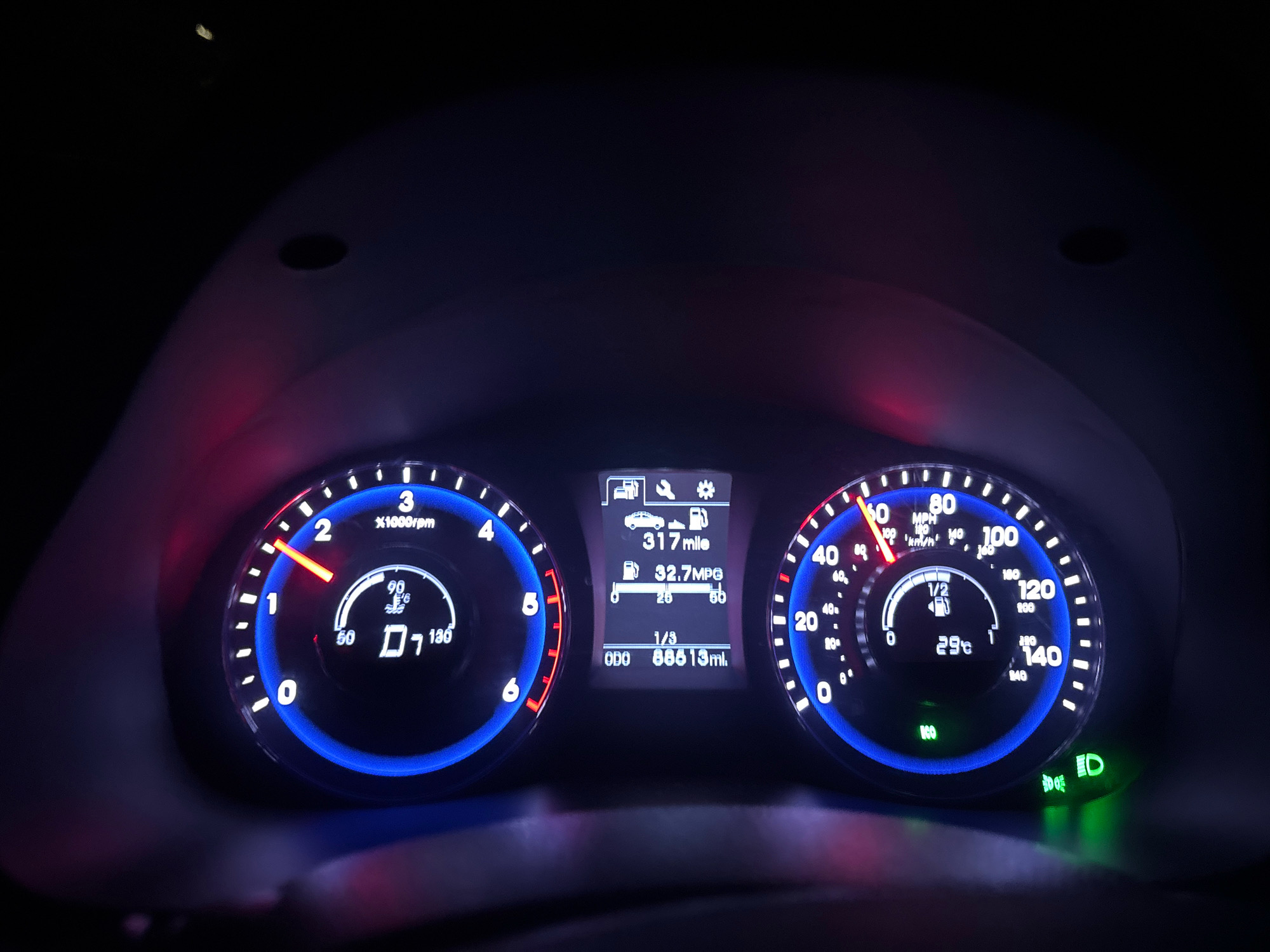 The image shows a close-up view of a car's dashboard at night, illuminated by the instrument cluster's lights. The dashboard features a tachometer on the left, a central digital display, and a speedometer on the right.
The tachometer shows the engine's revolutions per minute (RPM), with a needle pointing between 1 and 2, indicating approximately 1,500 RPM. It also displays the gear selection as "D" (Drive).
The central digital display shows various information:
- "317 mile": likely the trip odometer reading.
- "32.7 MPG": the fuel efficiency in miles per gallon.
- "1/3": possibly indicates the fuel level, with one-third of the tank remaining.
- "ODO 88813 mi.": the total mileage of the vehicle.
The speedometer on the right indicates a speed of approximately 50 MPH (80 km/h). It also shows the engine coolant temperature as 29°C. A green indicator light at the bottom right signifies the headlights are on.
The background is dark, suggesting it is nighttime. The interior of the car is dimly lit, with the bright blue and white lights of the dashboard being the primary light source. The image captures the operational status of a vehicle in motion or recently stopped. The specific location of Agios Tychona Community, Cyprus, is not discernible from the visual information presented in the image itself.