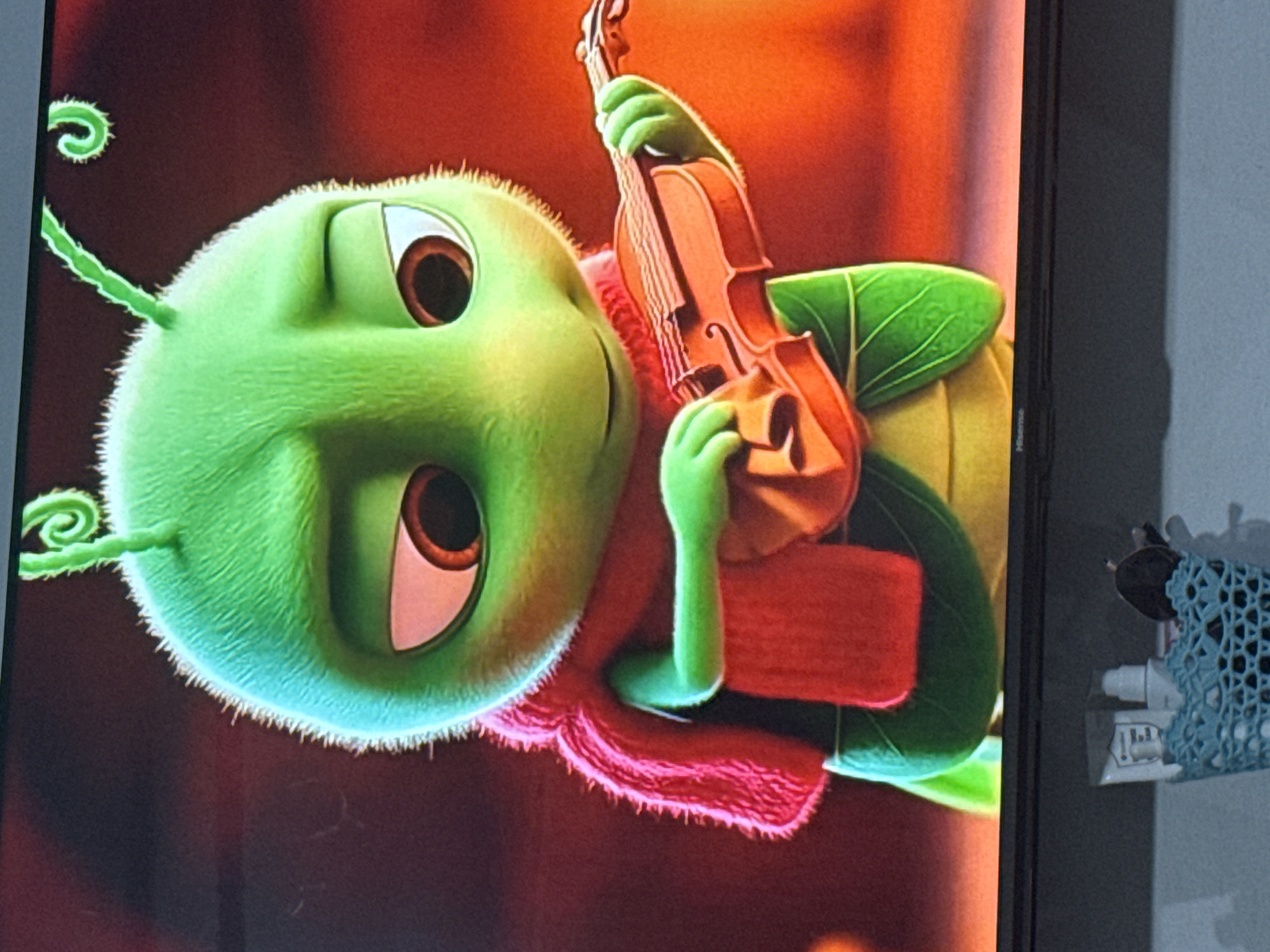 The image is a close-up shot of a cartoon caterpillar holding and playing a violin. The caterpillar is bright green with large, sad-looking eyes and curly antennae. It is wearing a red scarf. The background is a warm, blurry red and orange. The image is displayed on a television screen, with the bottom edge of the screen and a shelf with some items below it visible. There is no indication of the location being Chardara, Uzbekistan in the image.