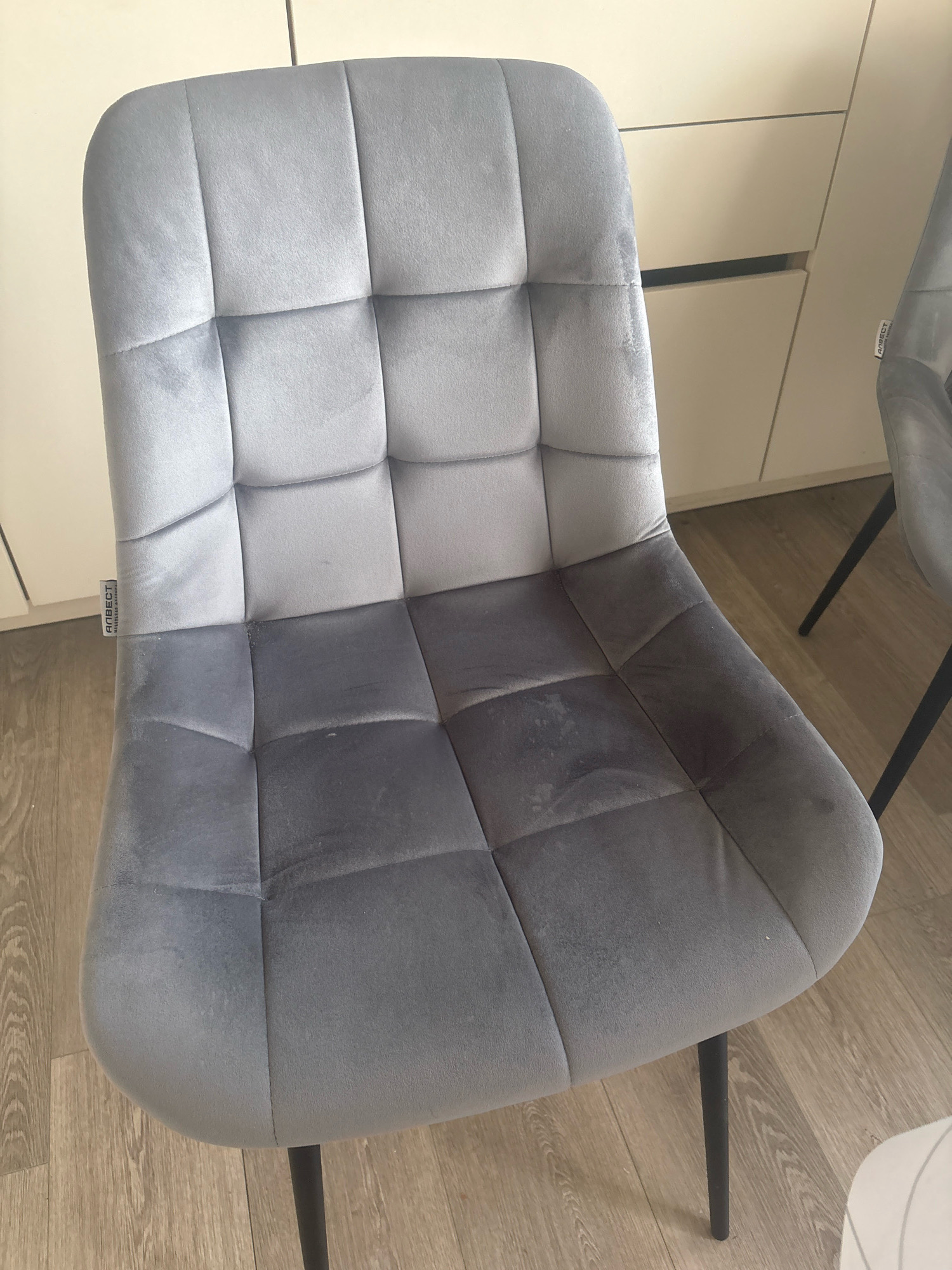 The image shows a close-up of a grey velvet chair with a tufted seat and back. The chair has black metal legs. To the left of the chair, there is a white cabinet or wall. To the right of the chair, another chair of the same design is partially visible, also with black metal legs. The flooring appears to be light-colored wood. A small white label with black text, likely a brand name, is attached to the left side of the chair. The scene is indoors and well-lit. There are no people or animals present in the image. There is no indication of the specific location within Dubovoye, Russia.