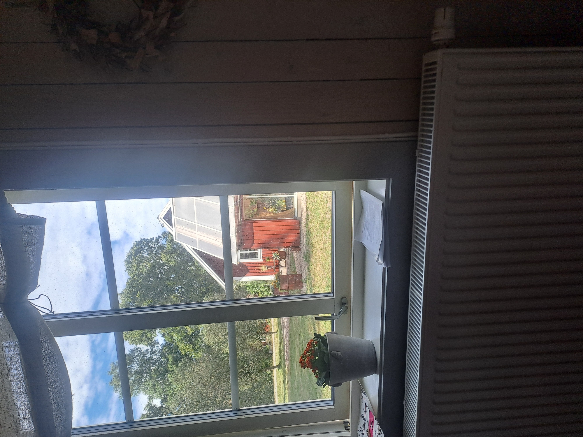 The image depicts an indoor scene looking out of a window in Björkvik, Sweden. In the foreground, a white radiator is visible below a windowsill. On the windowsill, a gray pot contains an orange flowering plant. Also on the windowsill are a few sheets of white paper. Through the window, a red building with a greenhouse attached is visible in a grassy yard surrounded by trees. A light blue sky with scattered clouds is seen above. To the right of the window, a wall paneled with vertical wooden planks is visible, with a wreath of dried leaves and twigs hanging on it. The overall impression is one of a rural or suburban setting in Sweden.