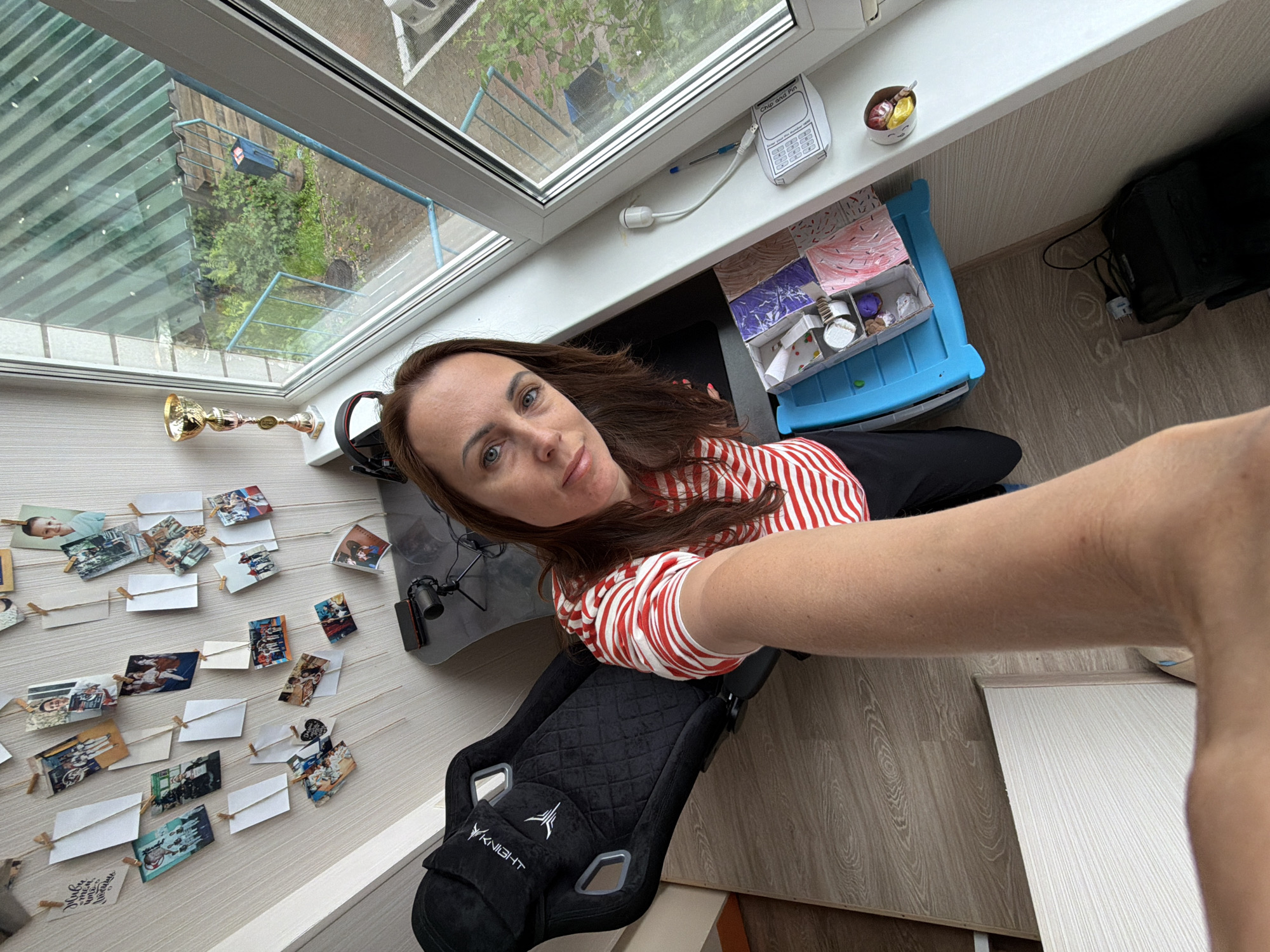 A Caucasian woman, estimated to be in her 30s to 40s, with long brown hair and blue eyes, is captured in a selfie-style image indoors. She is wearing a red and white striped short-sleeved top and dark pants. Her arm is extended towards the camera, suggesting she is holding the device.
The scene depicts an interior room, likely a residence, in the city of Birobidzhan, Russia. The room features light-colored, vertically striped wallpaper on the left wall and a light wood laminate floor. A large white-framed window dominates the right side of the image, offering a view of an urban or suburban landscape.
The left wall is decorated with multiple small photographs and blank white paper rectangles, arranged vertically along strings or wires and secured with miniature wooden clothespins. The photographs depict individuals, including children and adults, in various poses, some appearing to be associated with dance or performance. One white paper strip displays text in Russian cursive: "Живи в кайф, люби всем сердцем."
To the left of the woman, a black gaming chair with "KNIGHT" text and a stylized logo (possibly a "V") on the headrest is visible. The chair's upholstery features a diamond-quilted pattern. Partially visible behind the woman is a dark, irregularly shaped desk with a small black desktop microphone on a tripod and a dark rectangular electronic device (possibly a smartphone or tablet).
The windowsill beneath the window displays several items: a golden trophy with a red and gold base on the far left, a white USB dongle or sensor plugged into the wall, a blue pen, and a piece of paper taped down, labeled "Chip and Pin" with a numerical keypad diagram. A small white bowl containing colorful candies (yellow and red) is also on the windowsill.
Below the windowsill, on the floor, is a light blue plastic multi-compartment container holding various small craft items, including rolled papers and colored shapes. Adjacent to this, a black bag or electronic device with associated cabling is situated on the floor.
The exterior view through the window reveals a sloped green corrugated metal roof immediately below, followed by green foliage, indicating trees and shrubs. A blue metal fence is visible, alongside a dark blue cylindrical trash receptacle mounted on a pole. The ground appears to be a mix of paved and vegetated areas, consistent with a residential building or courtyard in an urban setting typical of Birobidzhan, Russia.