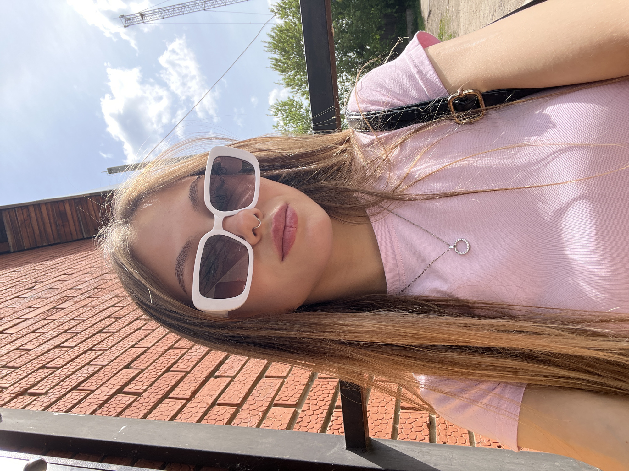 A young woman wearing white-framed sunglasses and a pink t-shirt poses for a selfie. She has long, light brown hair and a nose ring. Behind her is a red brick wall, and a crane is visible in the sky. The location is Фоки, Russia.