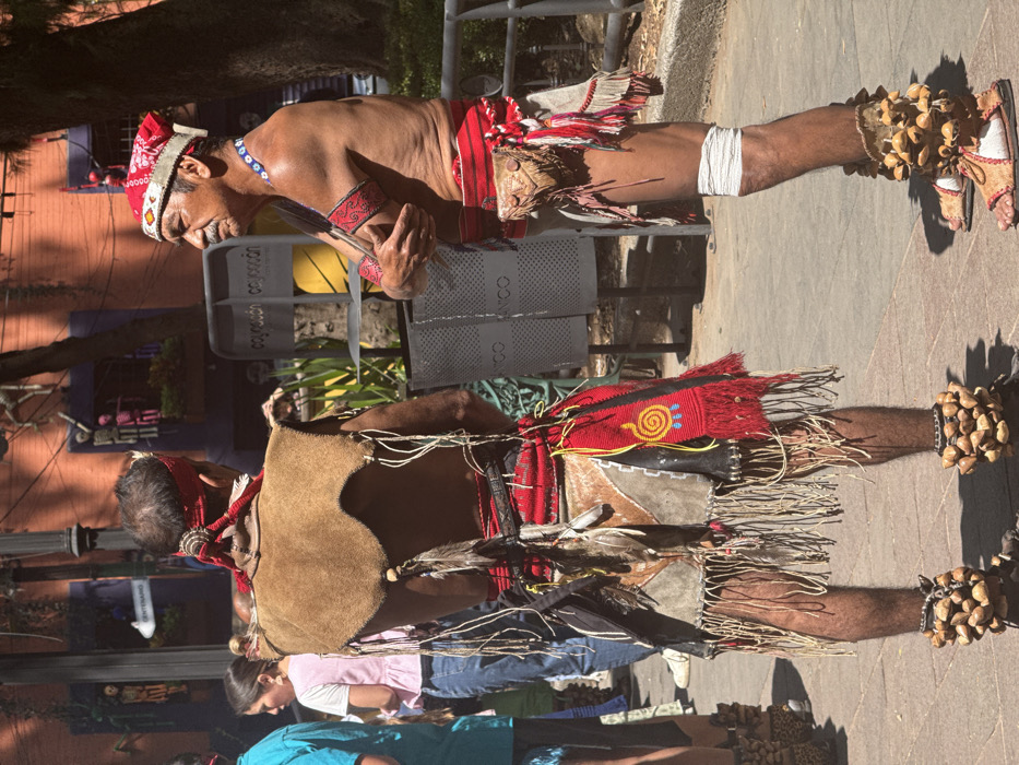 Two adult males in traditional Indigenous attire are depicted outdoors in the Coyoacán borough of Mexico City, Mexico, under direct sunlight. The man on the left, viewed from behind, wears a tan leather sleeveless top over a bare torso, a red headband, and a complex waist covering featuring red fabric, black leather, feathers, and fringes. His lower legs are covered in fringed material, and his ankles display large, round seed or nut rattles. The man on the right, viewed from his left side, is bare-chested with a mustache, wearing a red and white patterned headband, a blue beaded necklace, a red armband, and a red fabric waist wrap with an attached leather pouch. His lower right leg is wrapped in white fabric, and his feet are adorned with sandals and similar seed or nut rattles. He stands with his arms crossed over his chest, facing the man on the left. In the background, a building with terracotta and purple walls features skeletal decorations, and a street sign points to "CENTENARIO." Two grey cylindrical trash bins with the word "COYOACÁN" are positioned between the men and the building. The ground is paved.