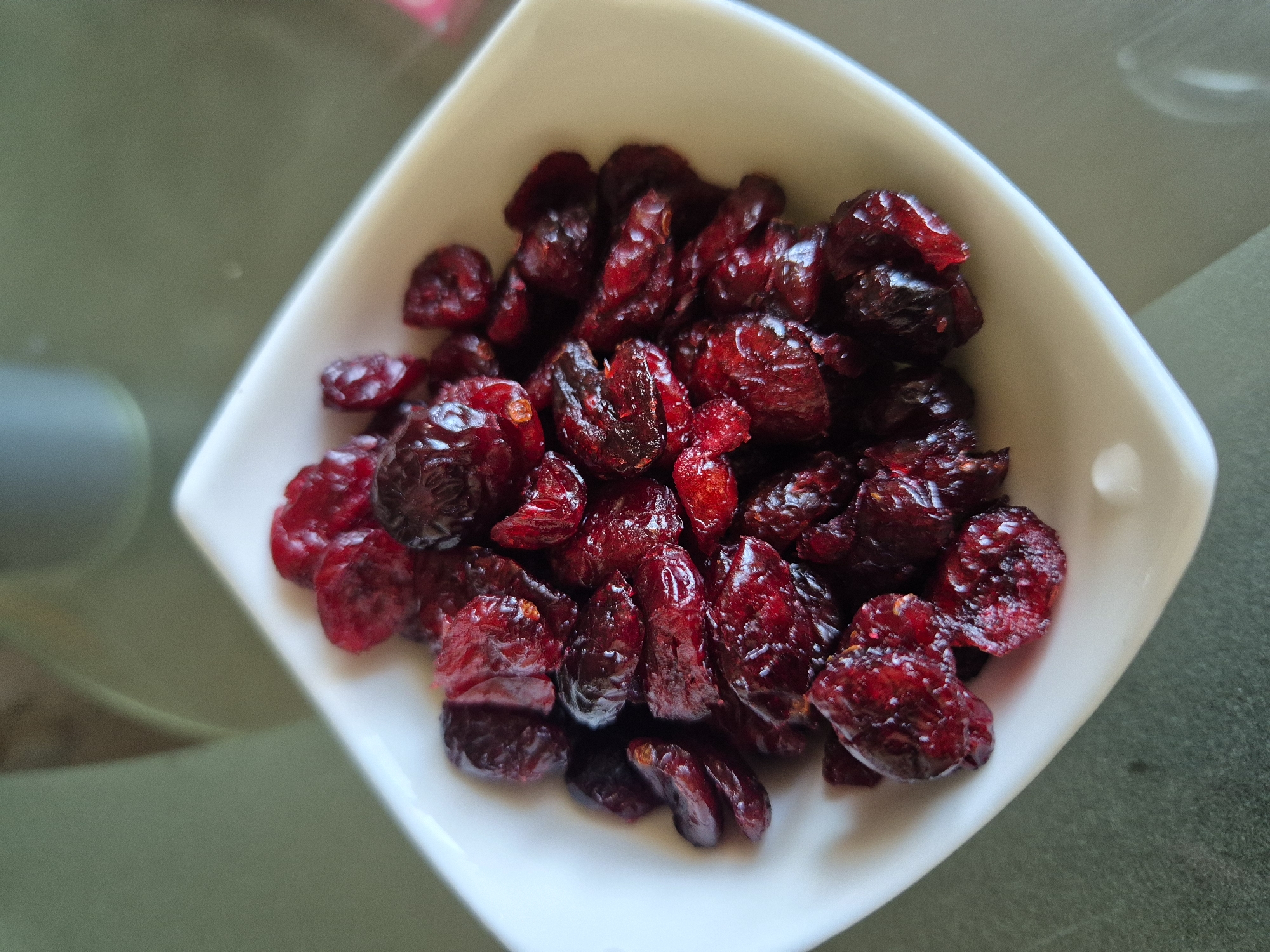 A close-up, top-down view shows a white, square bowl filled with dried cranberries. The cranberries are a deep red, almost maroon color, and are piled high in the bowl. Some appear to be whole, while others are broken or halved. The surface of the cranberries has a slightly glossy, wrinkled texture. The bowl is positioned on a dark, speckled surface that appears to be a tabletop. The background is softly blurred, with hints of green and gray, suggesting an indoor setting. There are no people visible in the image.