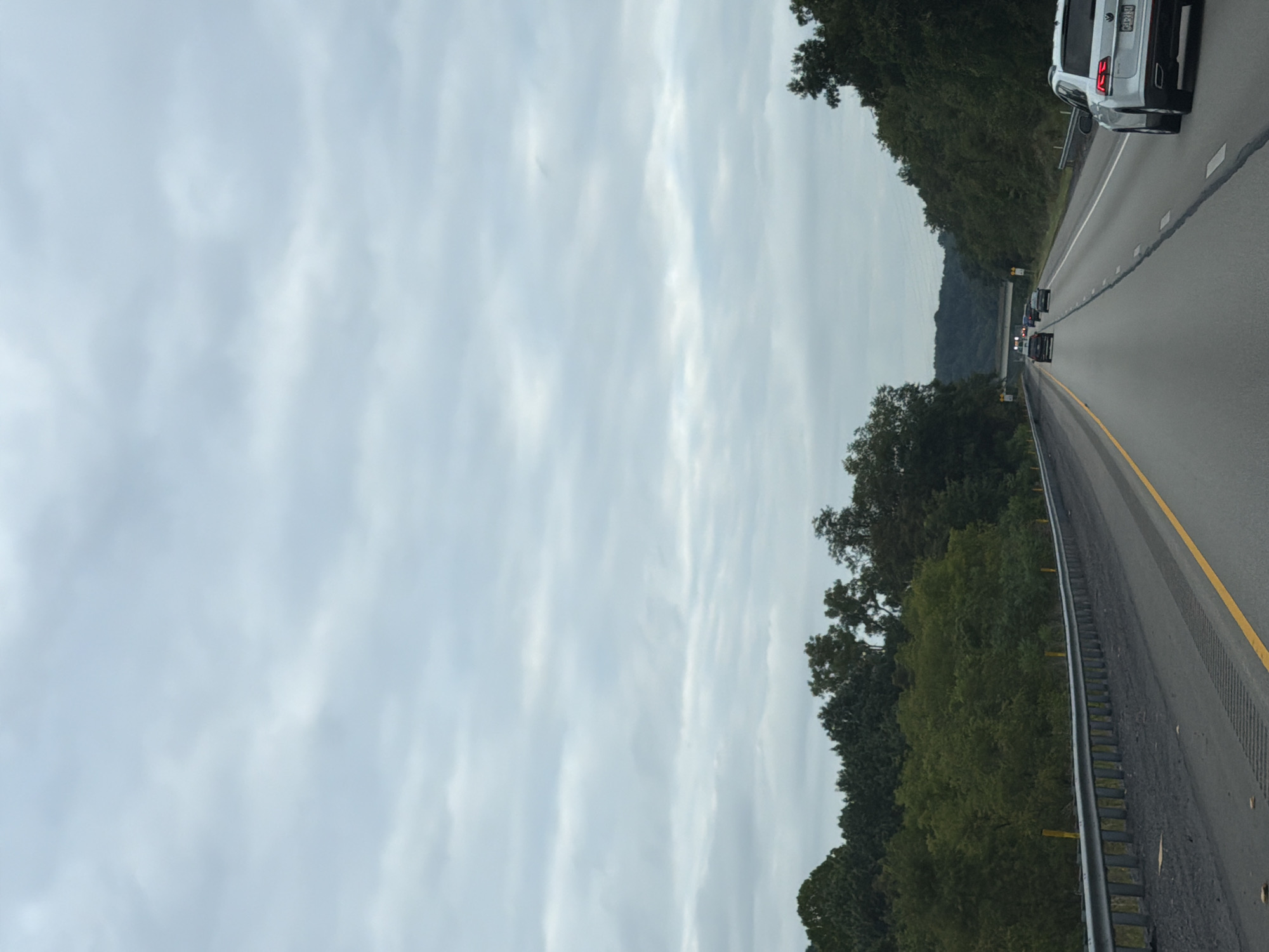 An overcast sky looms over a highway in Kennedy Township, United States of America. The highway has multiple lanes of asphalt with yellow lane markers. Guardrails line the sides of the road, with trees and foliage growing behind them. Several cars are driving on the highway. One white SUV is in the foreground on the right side of the image, while other vehicles are further down the road. A bridge spans across the highway in the distance.