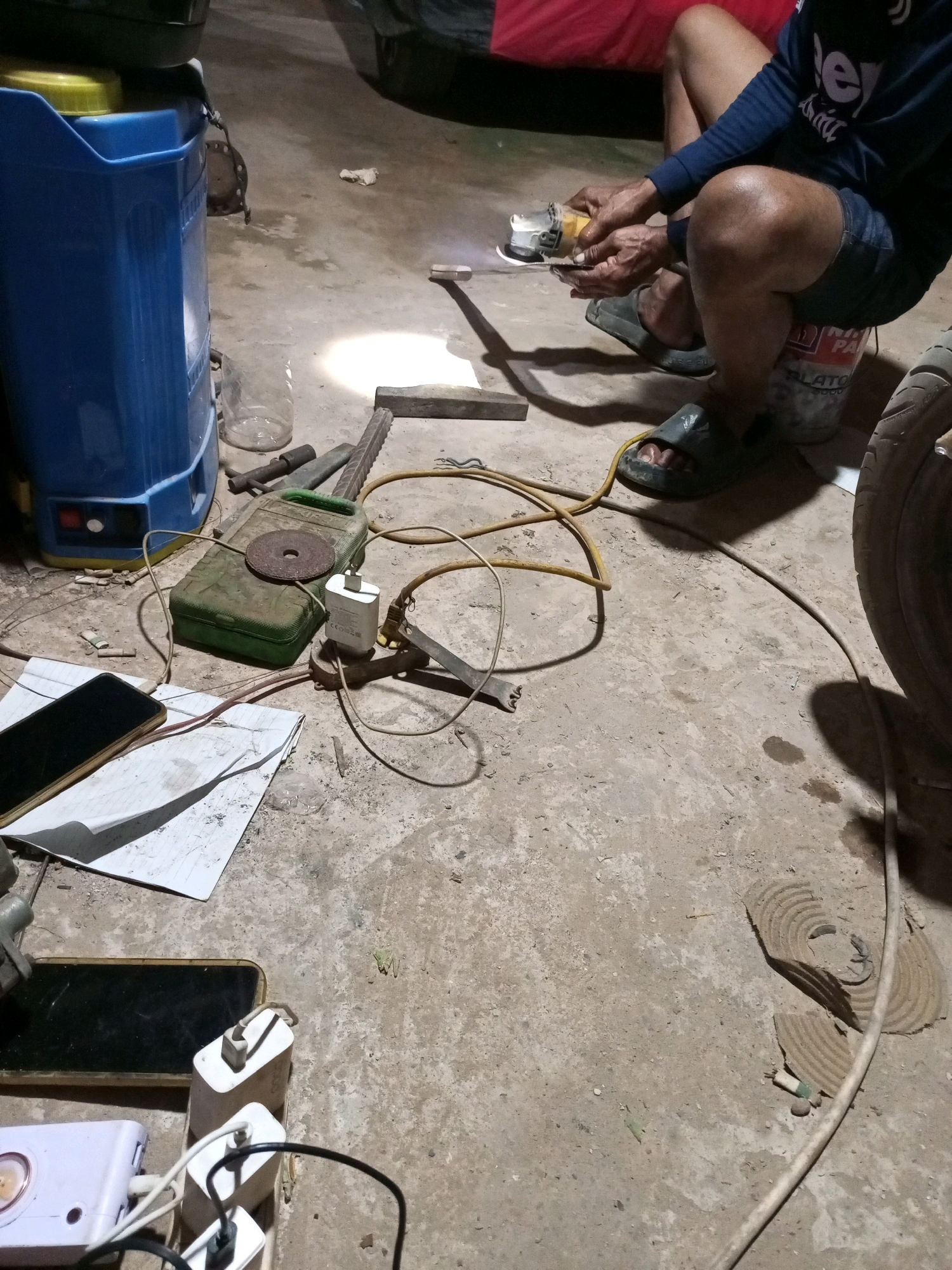 A person is working on a metal object with a grinder in an outdoor setting, possibly a garage or workshop. The ground is concrete, and there are various tools and electrical equipment scattered around. A large blue plastic container is to the left of the scene, and a red cloth is visible in the background. The lighting suggests it may be nighttime or a dimly lit area.